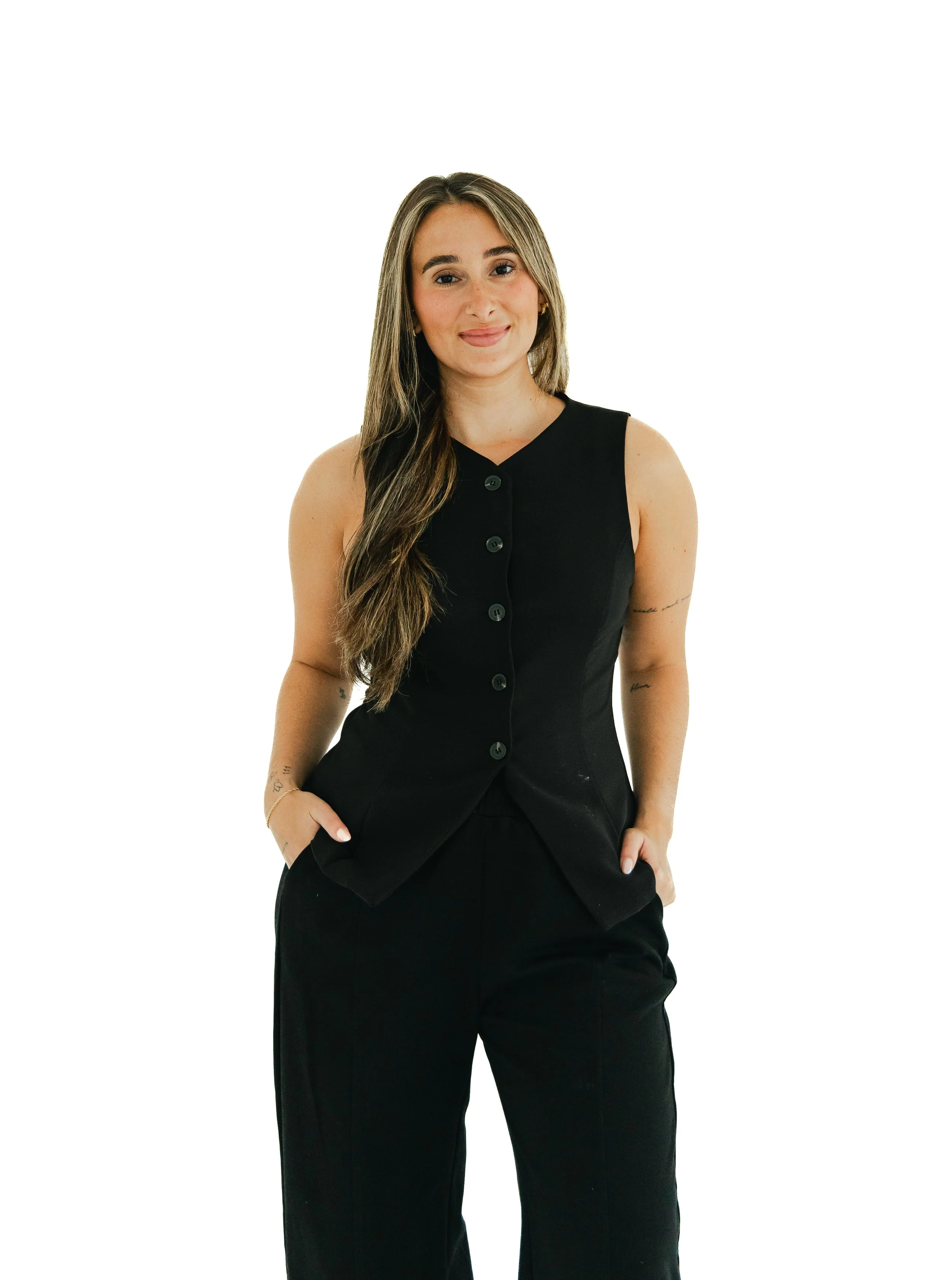 A young woman with long wavy hair, wearing a sleeveless black button-up top and black pants, standing against a plain white background.