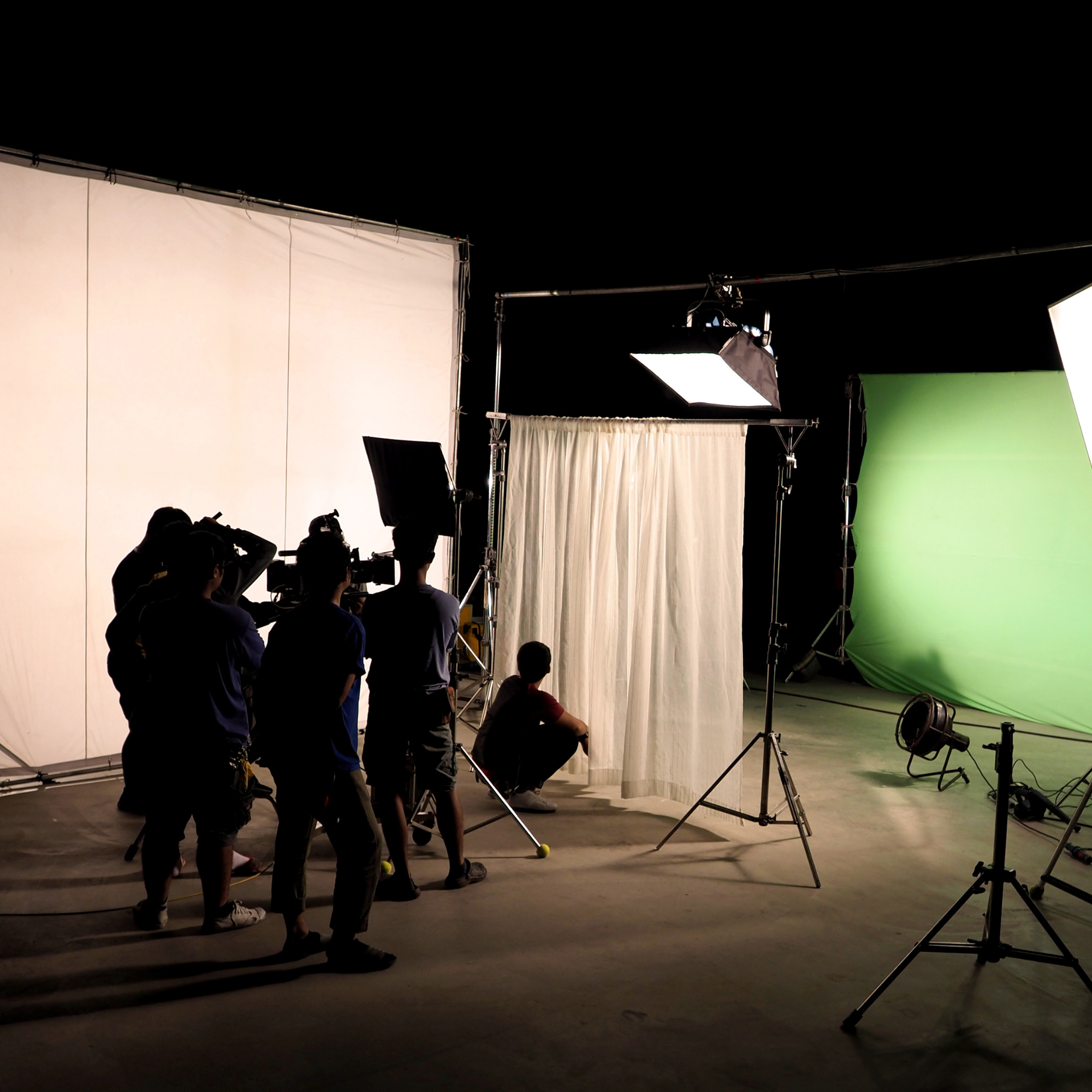 Behind-the-scenes of a film or photo shoot with lighting equipment, a green screen, a white backdrop, and several crew members working in a studio.