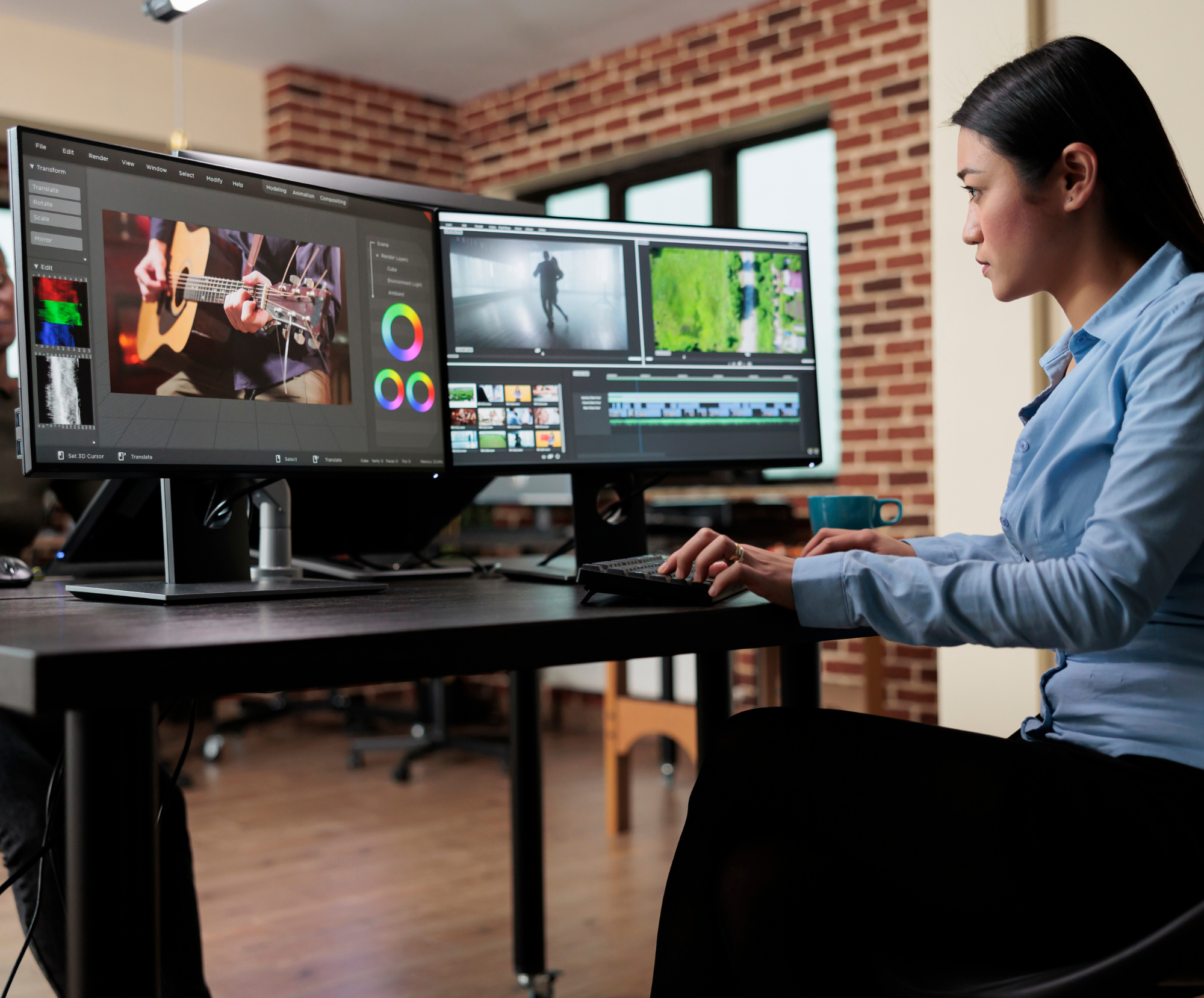 A woman with long black hair working on video editing on two computer monitors in a room with brick walls and large windows.