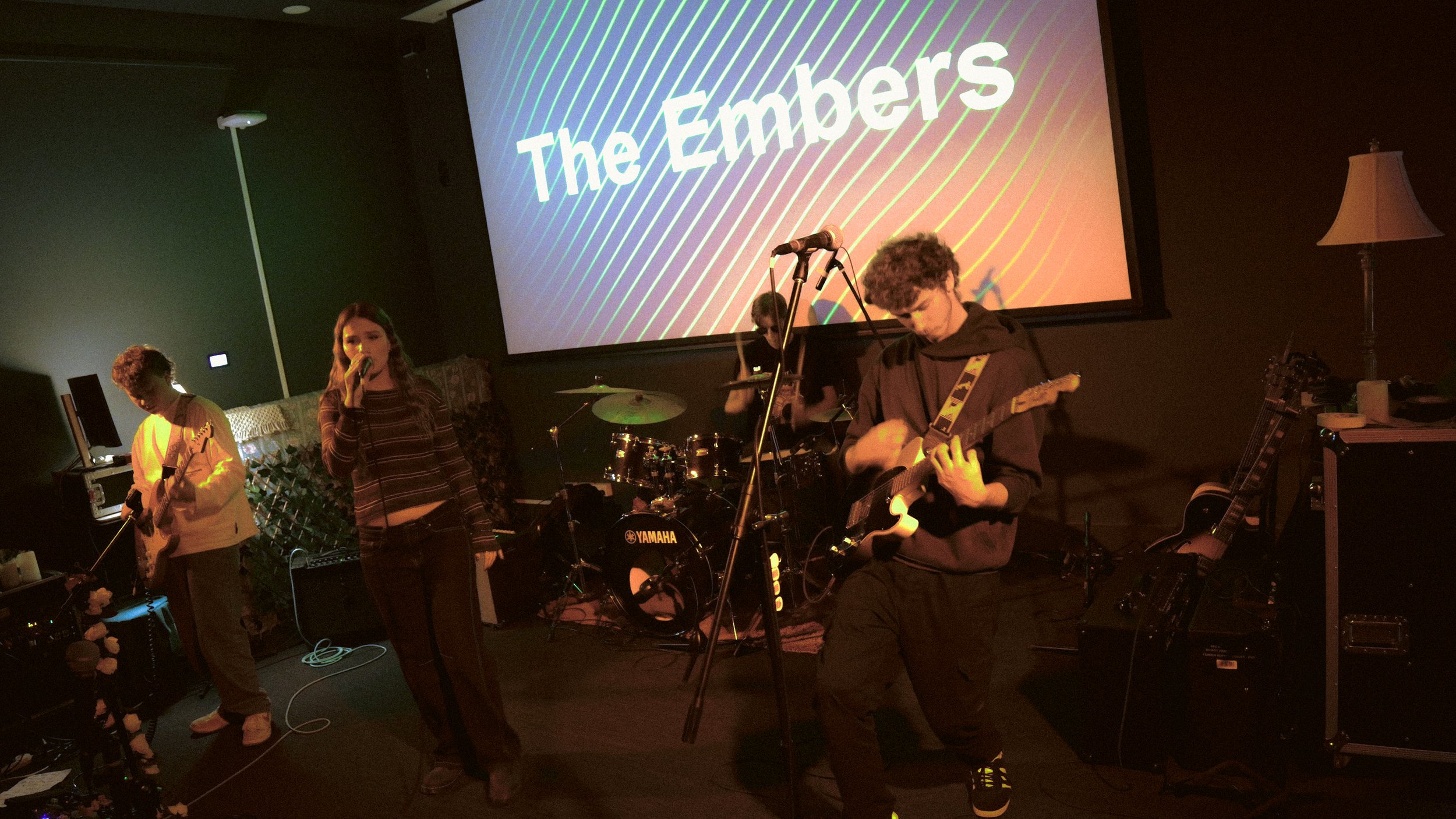 A band performing on stage with two guitarists, a singer, and a drummer, with a large screen behind them displaying the text 'The Embers'.