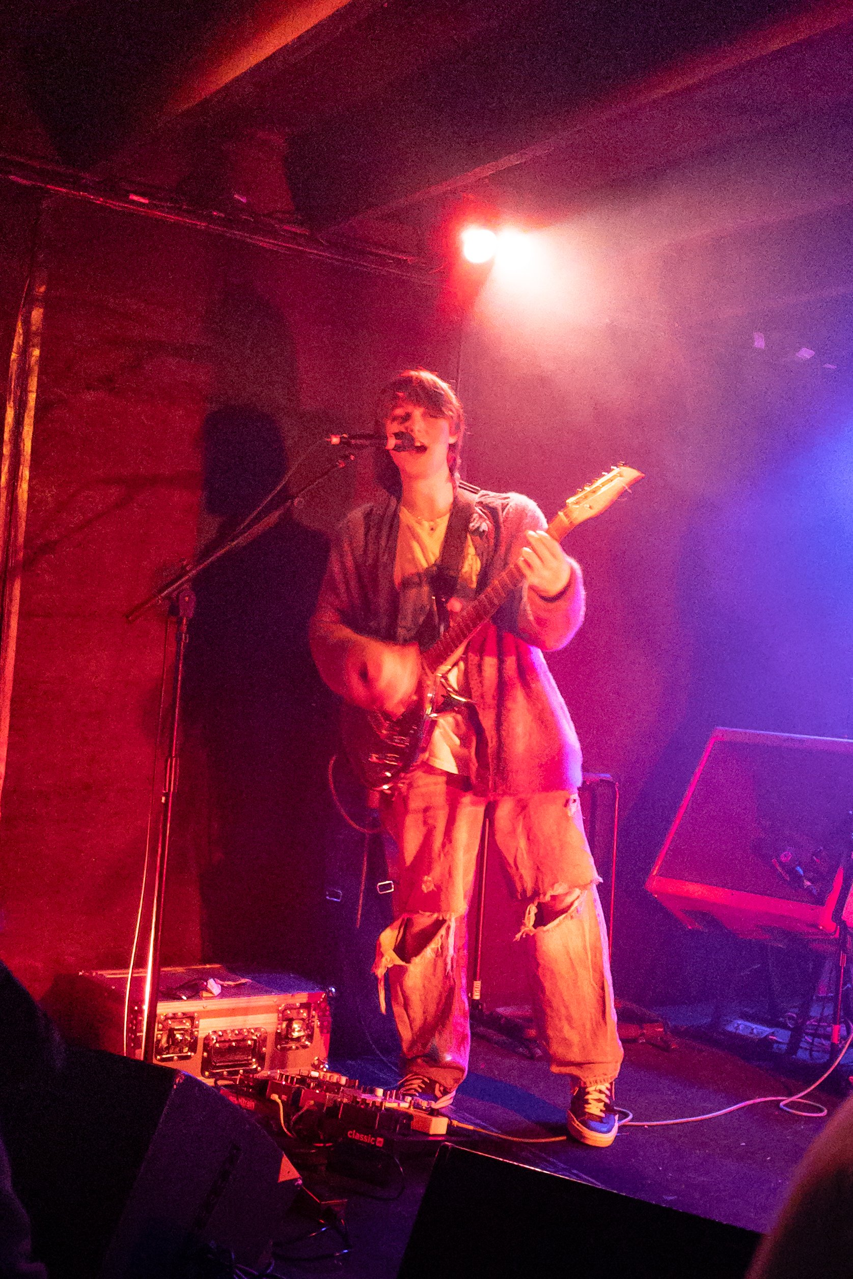 Musician performing on stage with guitar, surrounded by colorful stage lighting and musical equipment.
