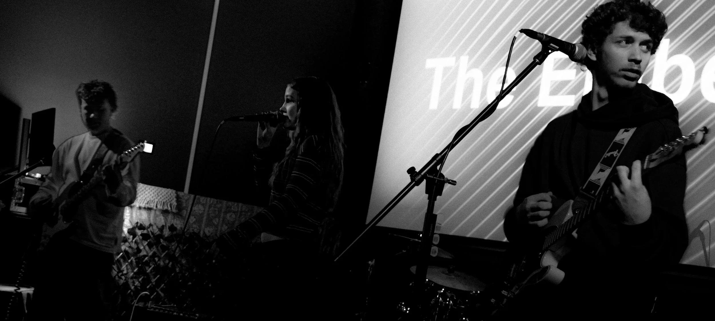 Three young musicians performing on stage with musical instruments, including guitars and microphone, in front of a large screen with the words 'The Echo' visible.