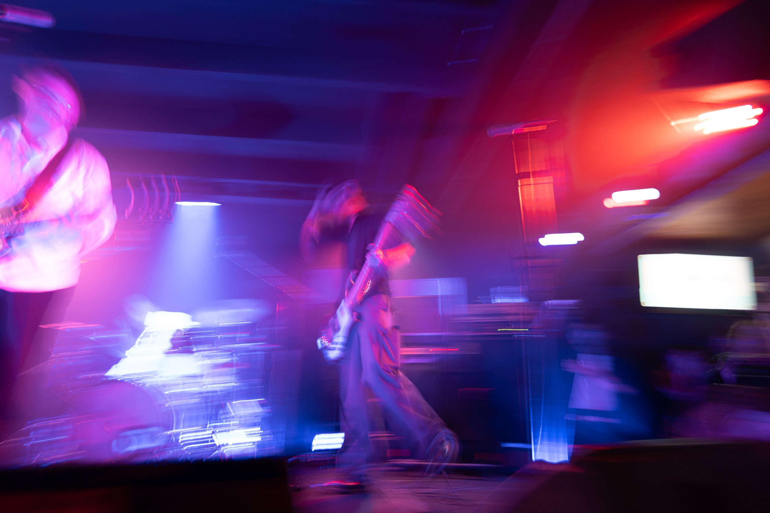 Blurred image of two people performing on stage with purple, pink, and red lighting.