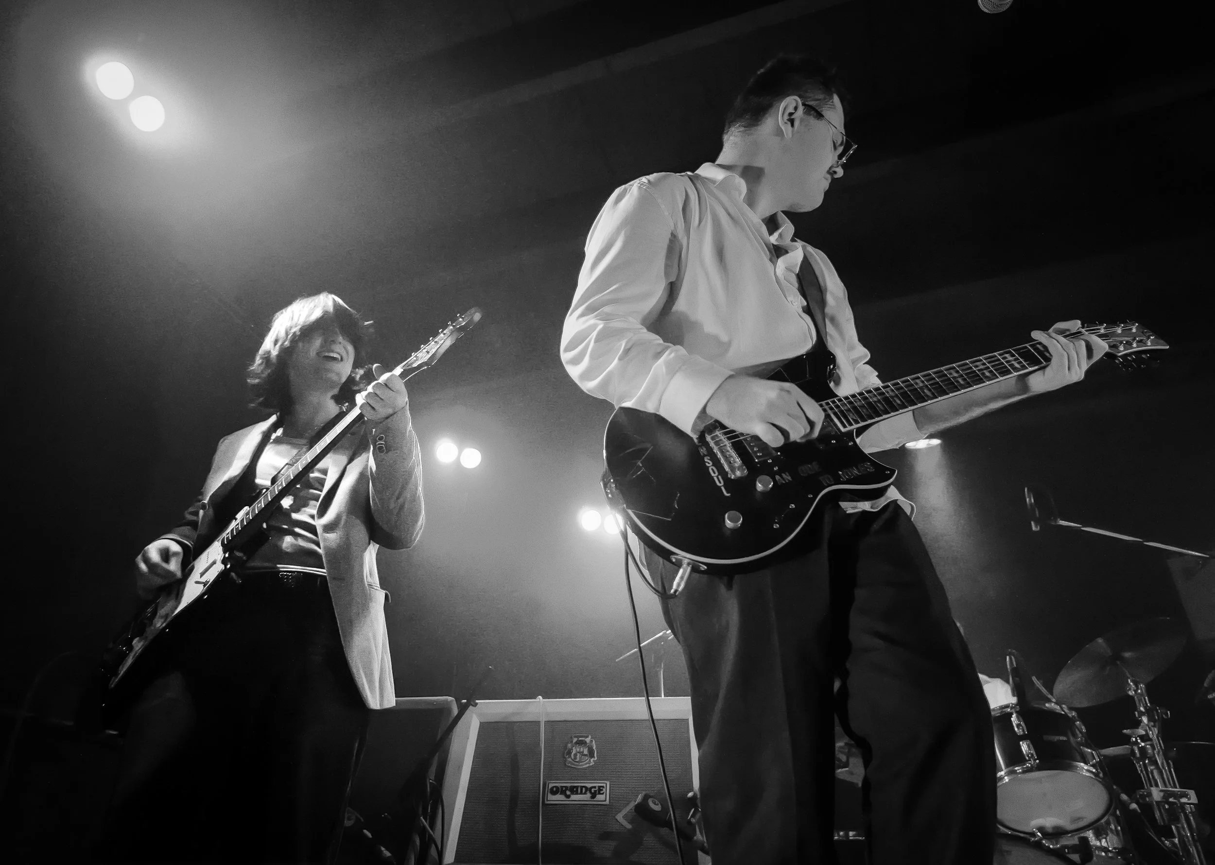 Two musicians playing electric guitars on stage under concert lighting, one woman smiling and another man focused on playing, with a drum set and amplifier in the background.