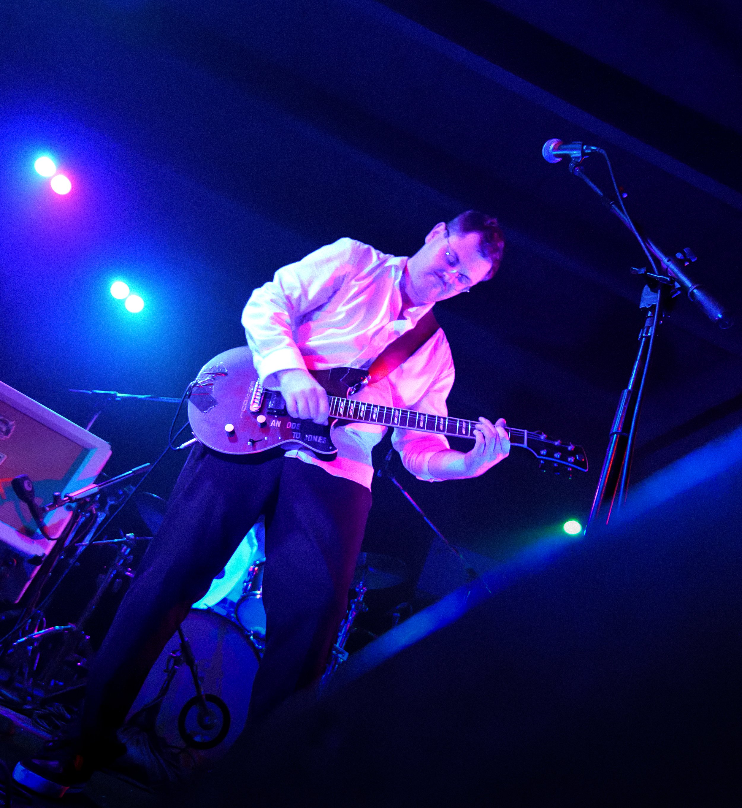 A musician playing an electric guitar on stage under blue and purple stage lights. The musician is wearing glasses and a light-colored shirt, with a microphone stand nearby.
