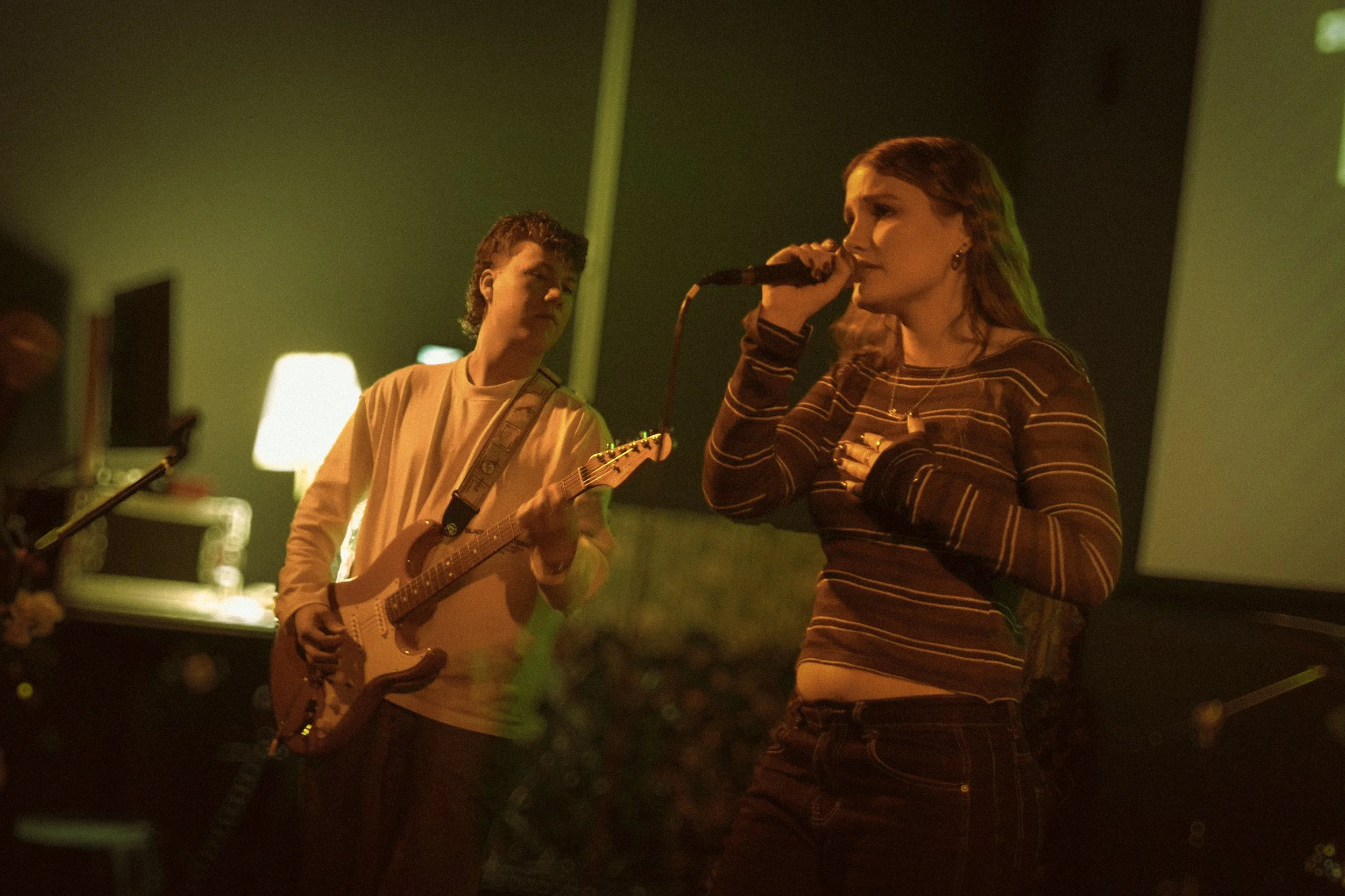 A woman singing into a microphone and a young man playing an acoustic guitar in a dimly lit room with green walls.