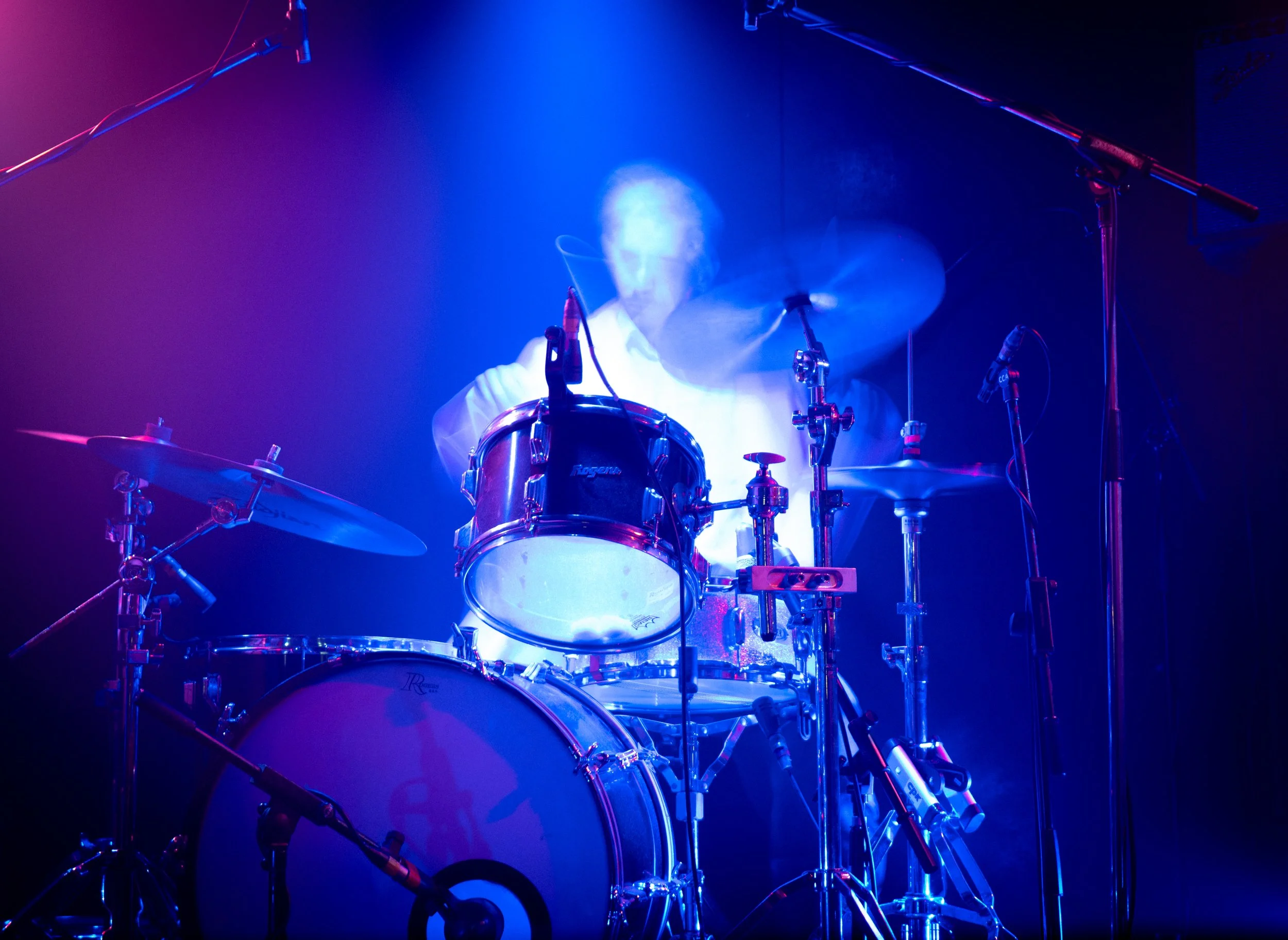 A drummer playing on stage illuminated by colorful purple and blue lights, with visible drums and microphones.