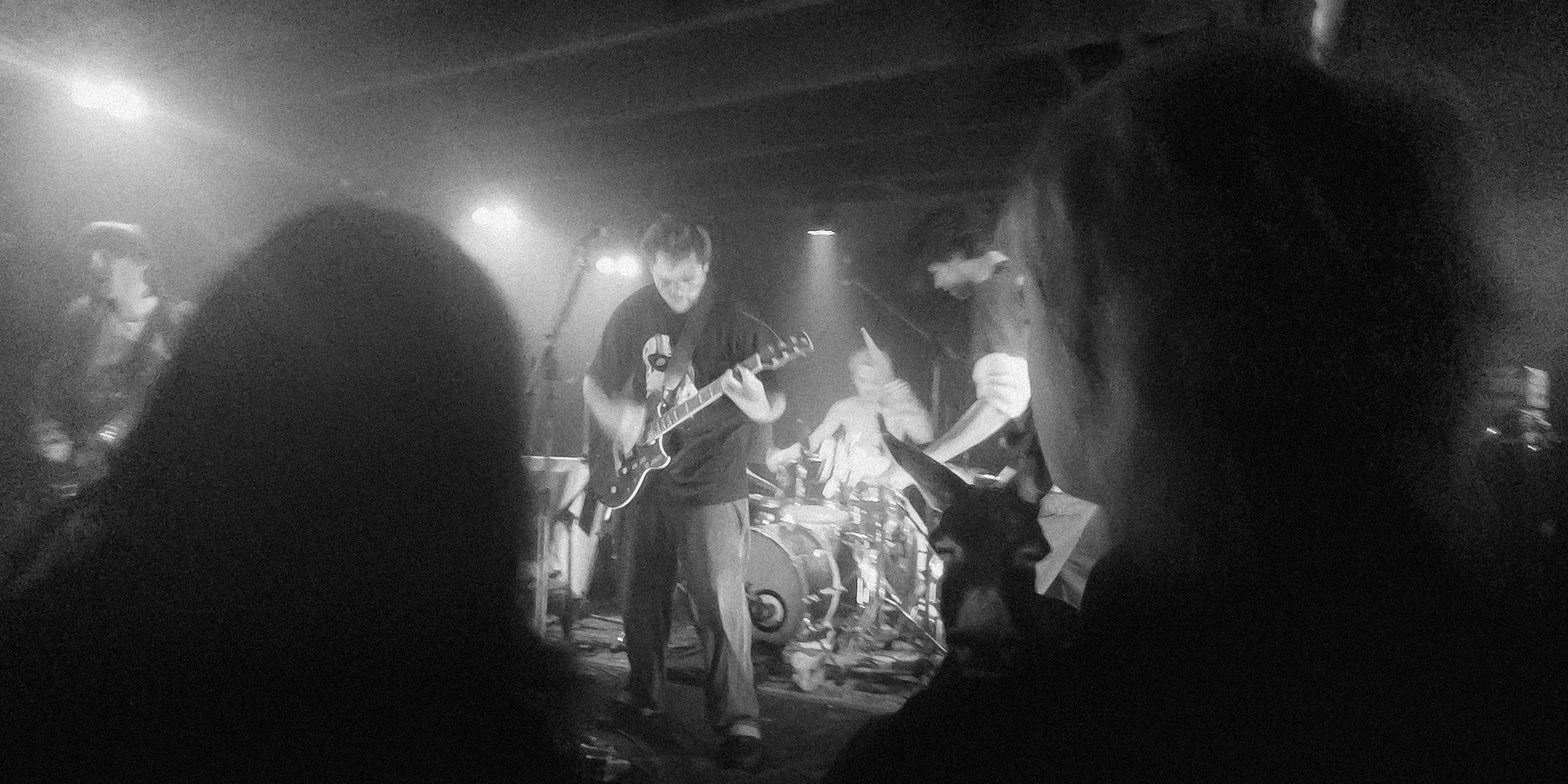 A black-and-white photo of a band performing live on stage, with several musicians playing instruments, and audience members in the foreground.