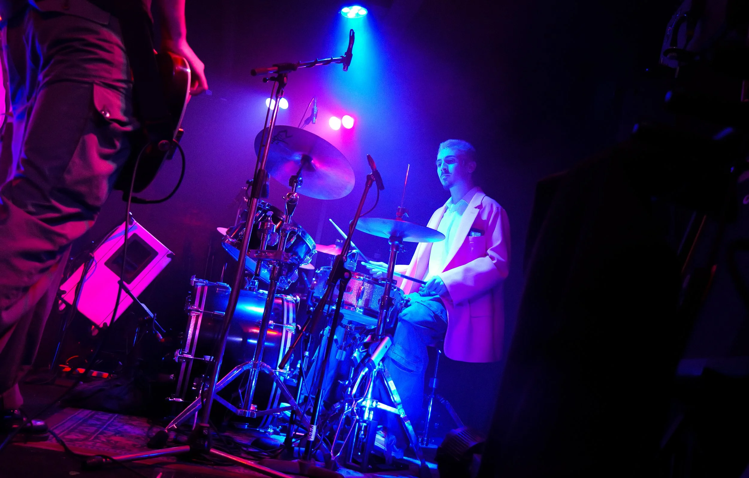 Young man in a white blazer playing drums on stage with colorful stage lighting.