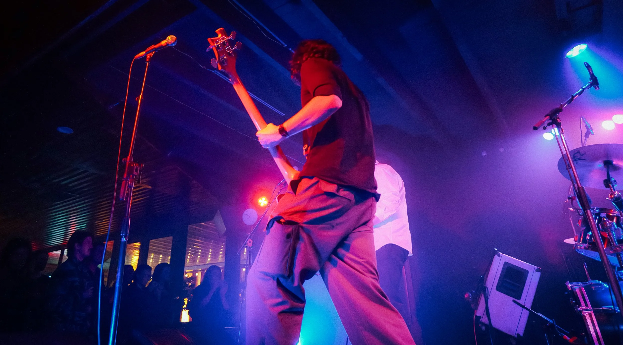 A musician playing an electric guitar on stage under colorful stage lights, with a crowd in the background.