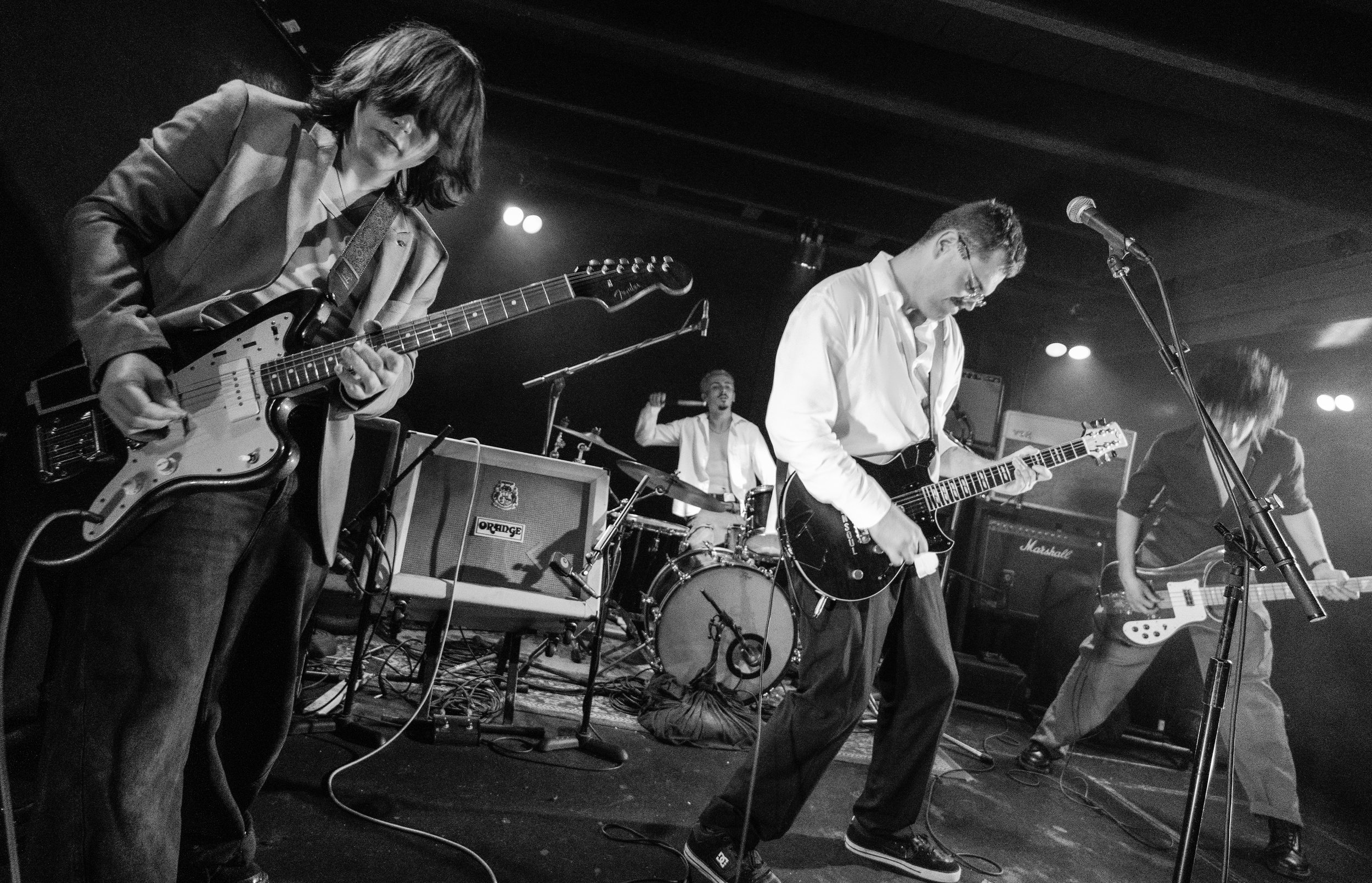 Black and white photo of a band performing on stage. There are several musicians, including guitarists and a drummer, with amplifiers and microphones. One guitarist is playing an electric guitar, while another is kneeling and playing an electric guitar. The drummer is in the background.