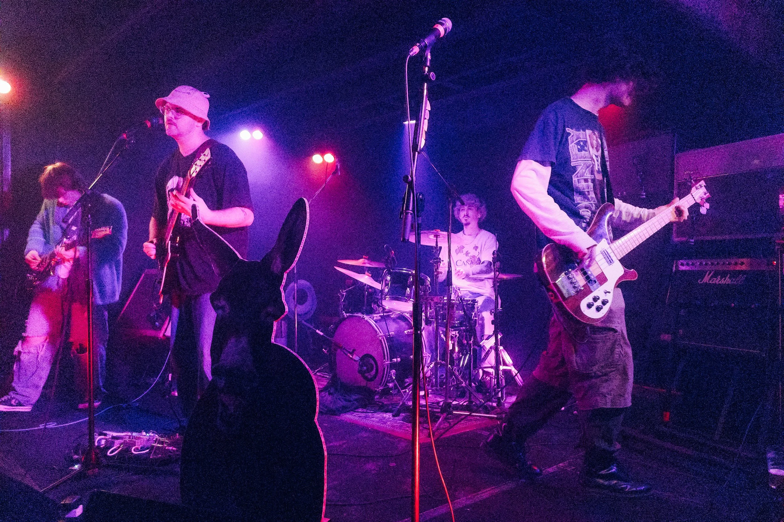 A band performing live on stage with purple and pink lighting, featuring guitarists, a bassist, and a drummer.