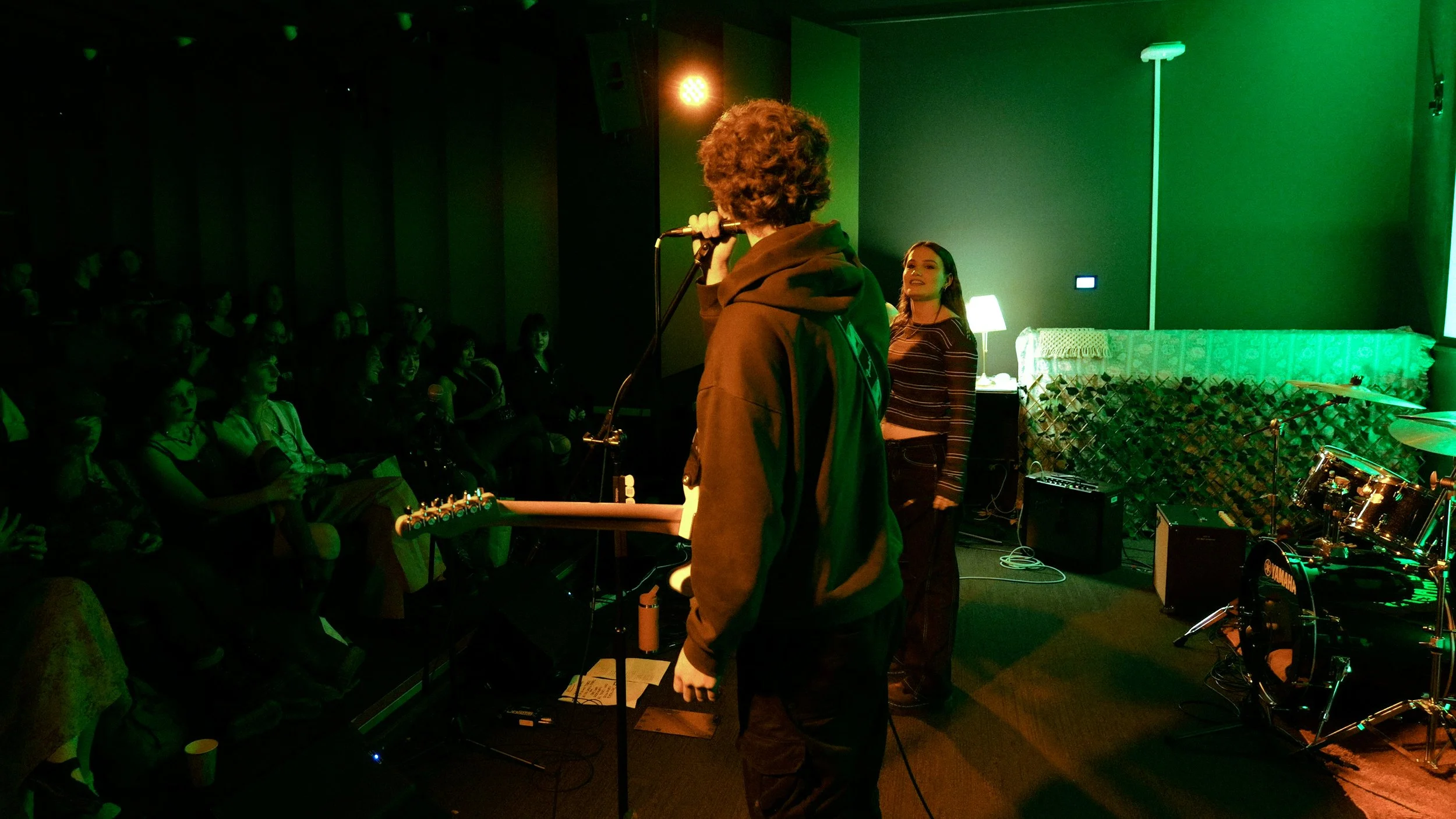A performer with curly hair and a hoodie standing on stage with a microphone, facing an audience in a dimly lit music venue with green and yellow lighting, musical instruments, and a woman in striped shirt smiling.
