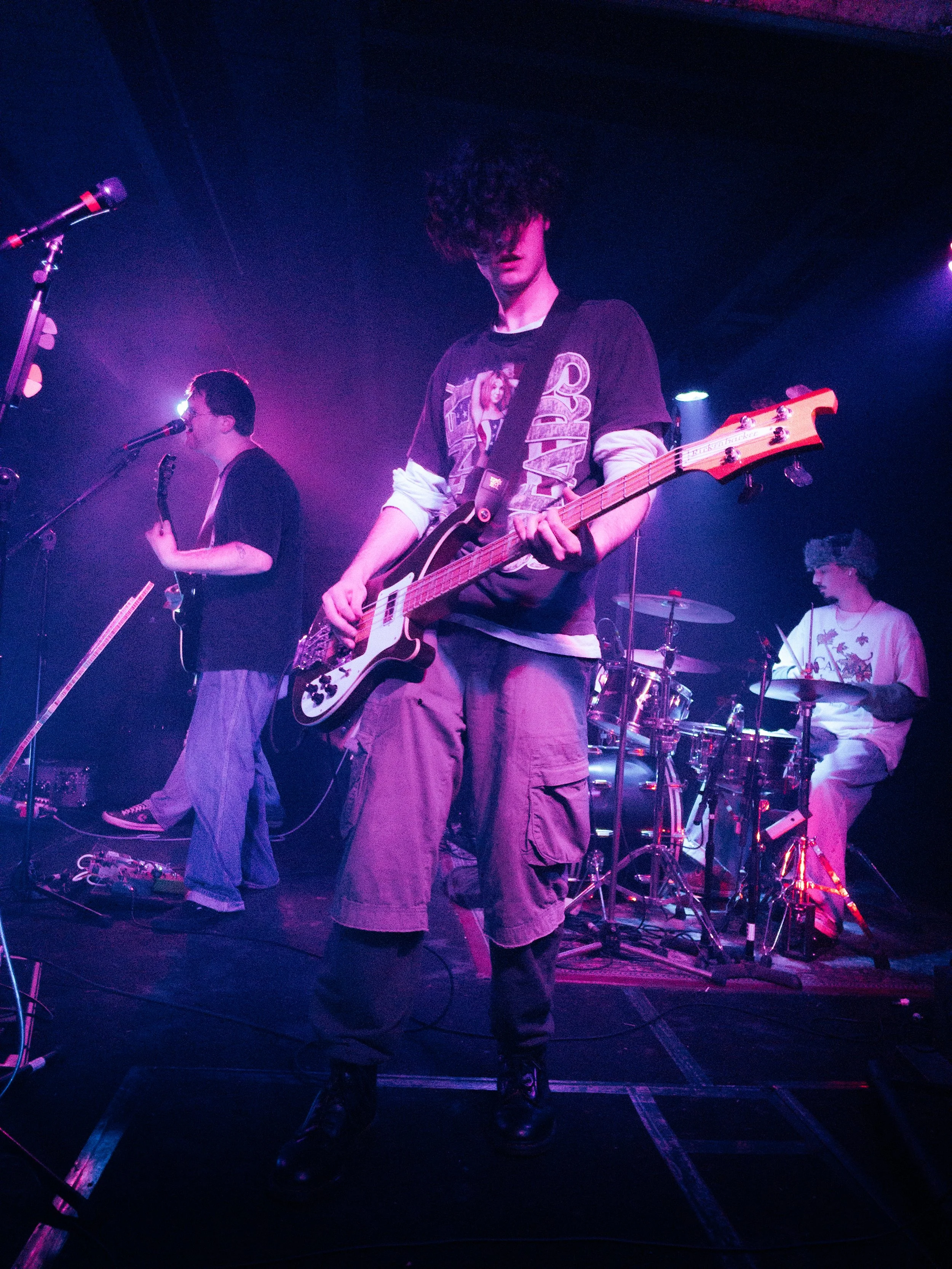 Three young musicians performing on stage with purple and pink lighting. One playing bass guitar, another singing into a microphone, and a third on drum set in the background.