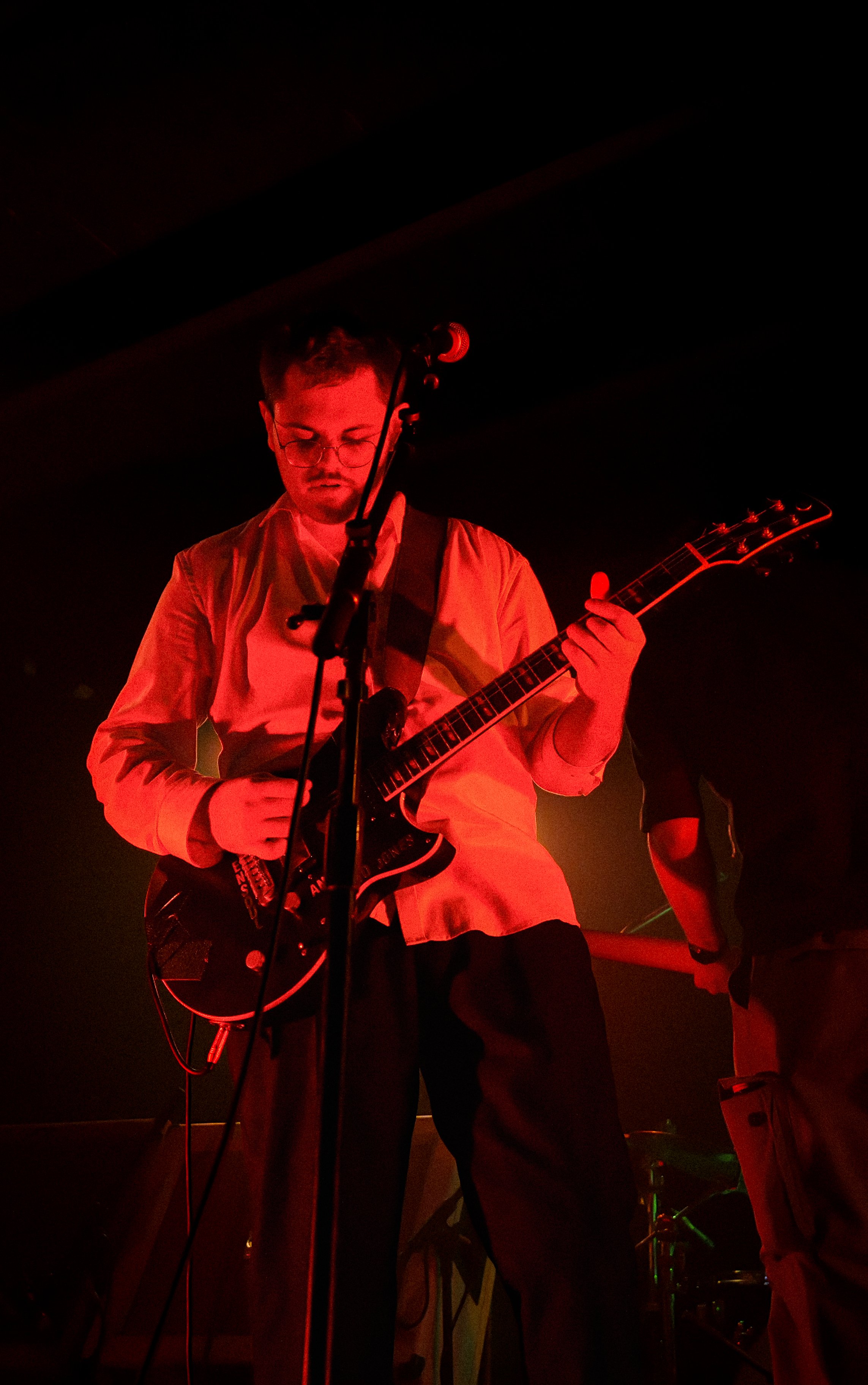 A man wearing glasses plays an electric guitar on stage illuminated by red lighting.