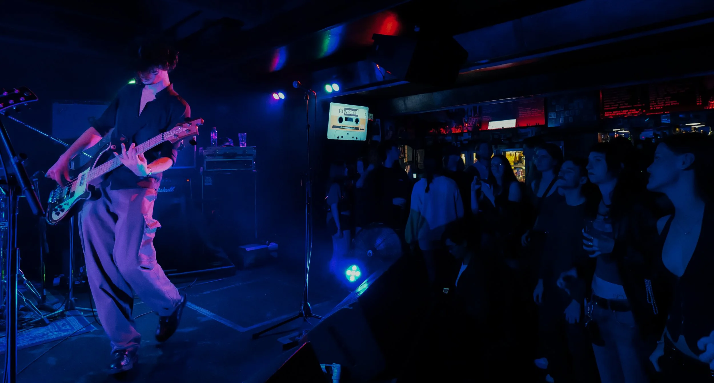 A musician playing an electric guitar on a dimly lit stage with blue and purple lights, and an audience watching him.