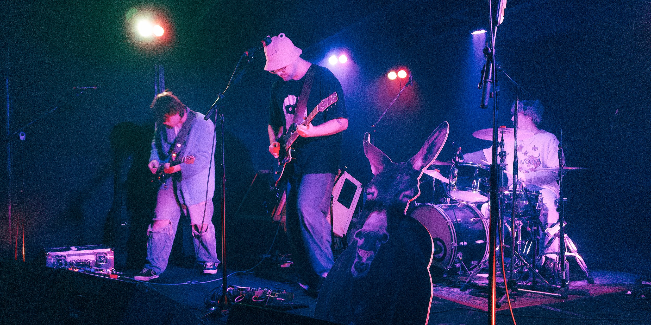 A band performing on stage with three members visible: a guitarist on the left, a bassist in the center, and a drummer on the right, illuminated by colorful stage lights.