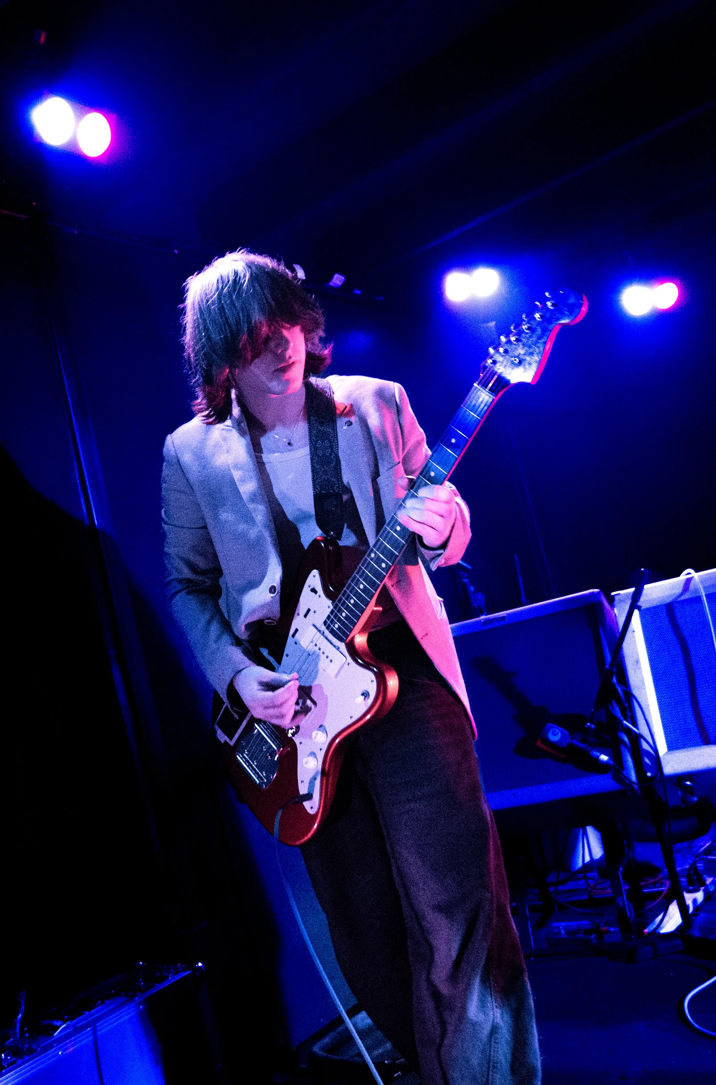 A young person with shoulder-length hair playing an electric guitar on stage, illuminated by blue and purple stage lights.