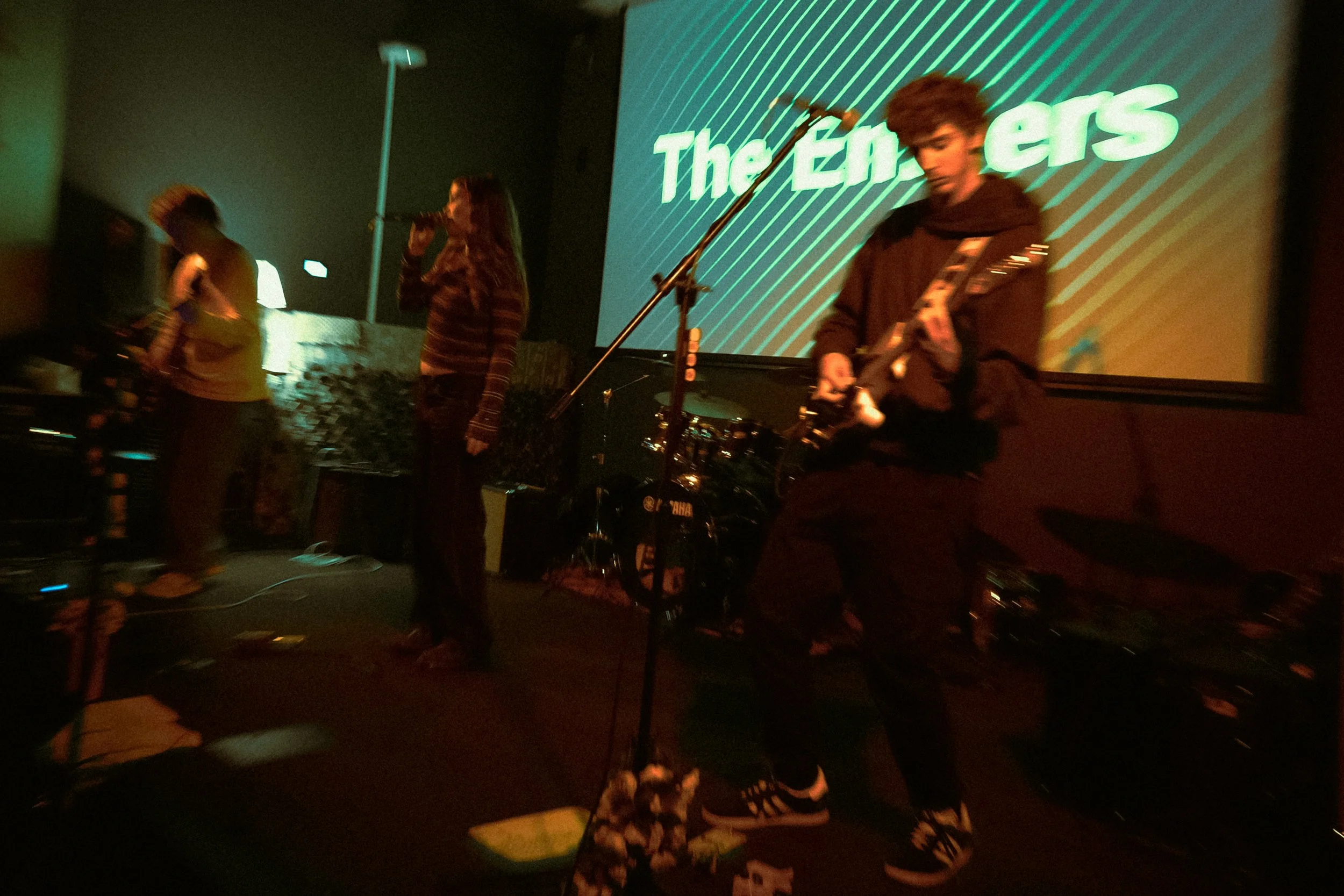 A band performing on stage with a large screen behind them displaying the words "The En" in green text. The scene is dark and the performers are illuminated by low stage lighting.