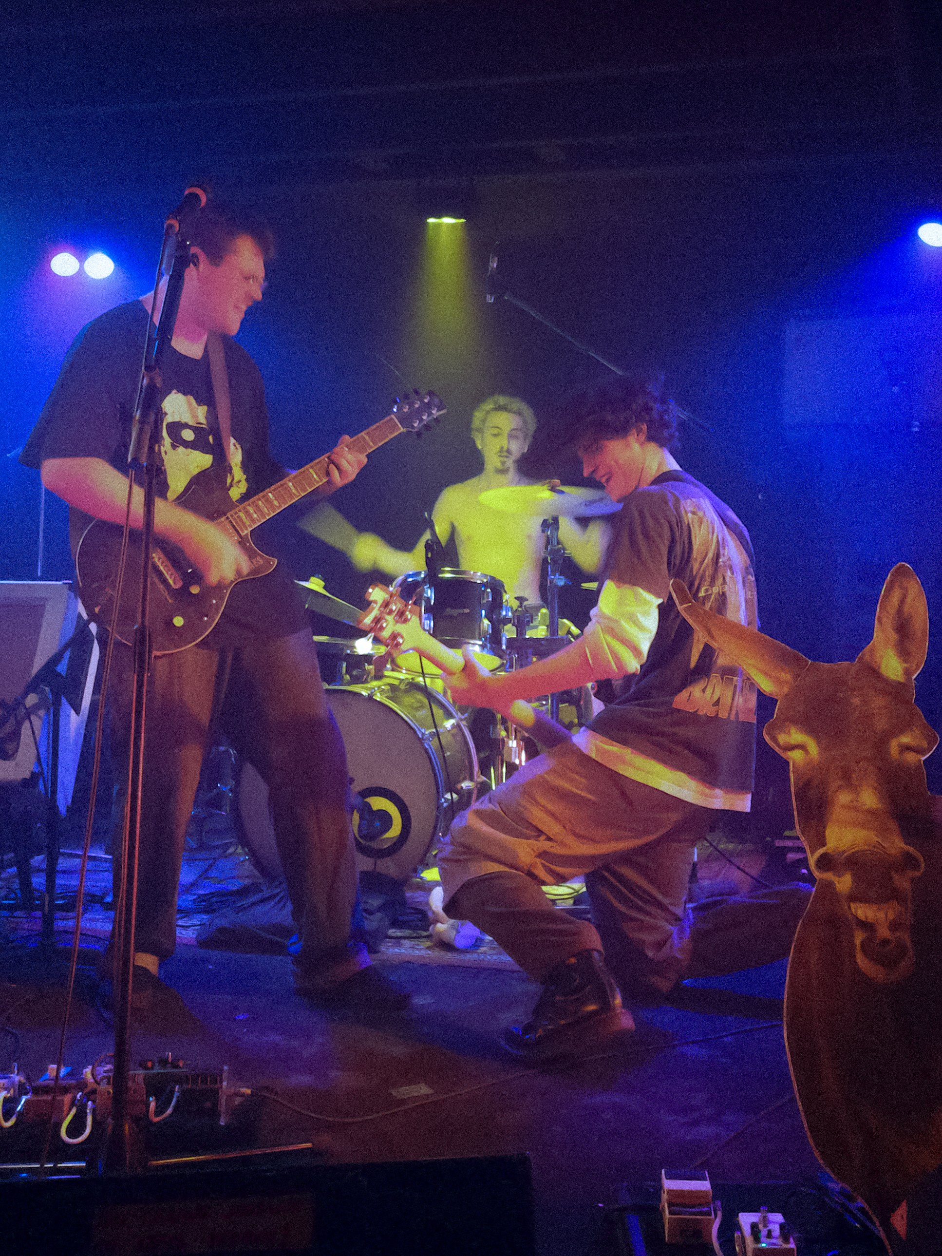 Three young men are performing on a dark stage under colorful lighting, playing guitars and drums with no audience visible.