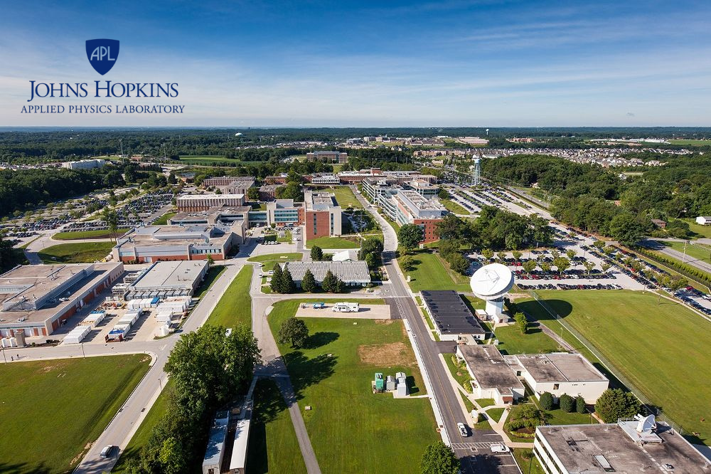 Aerial view of Johns Hopkins Applied Physics Laboratory campus with buildings, parking lots, green lawns, and a satellite dish antenna under a blue sky.
