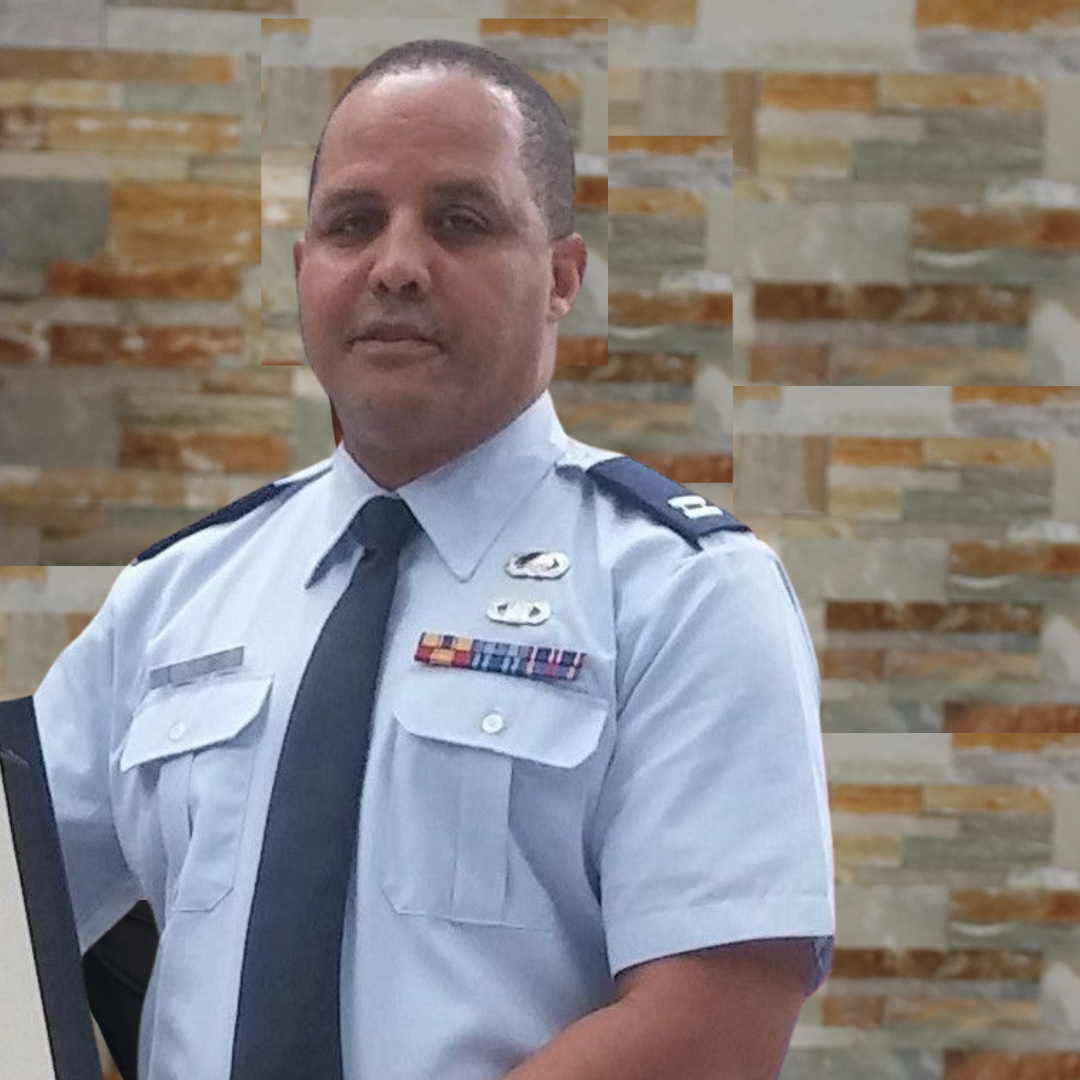 A man in a light blue military uniform with medals and patches, standing in front of a multicolored brick wall.