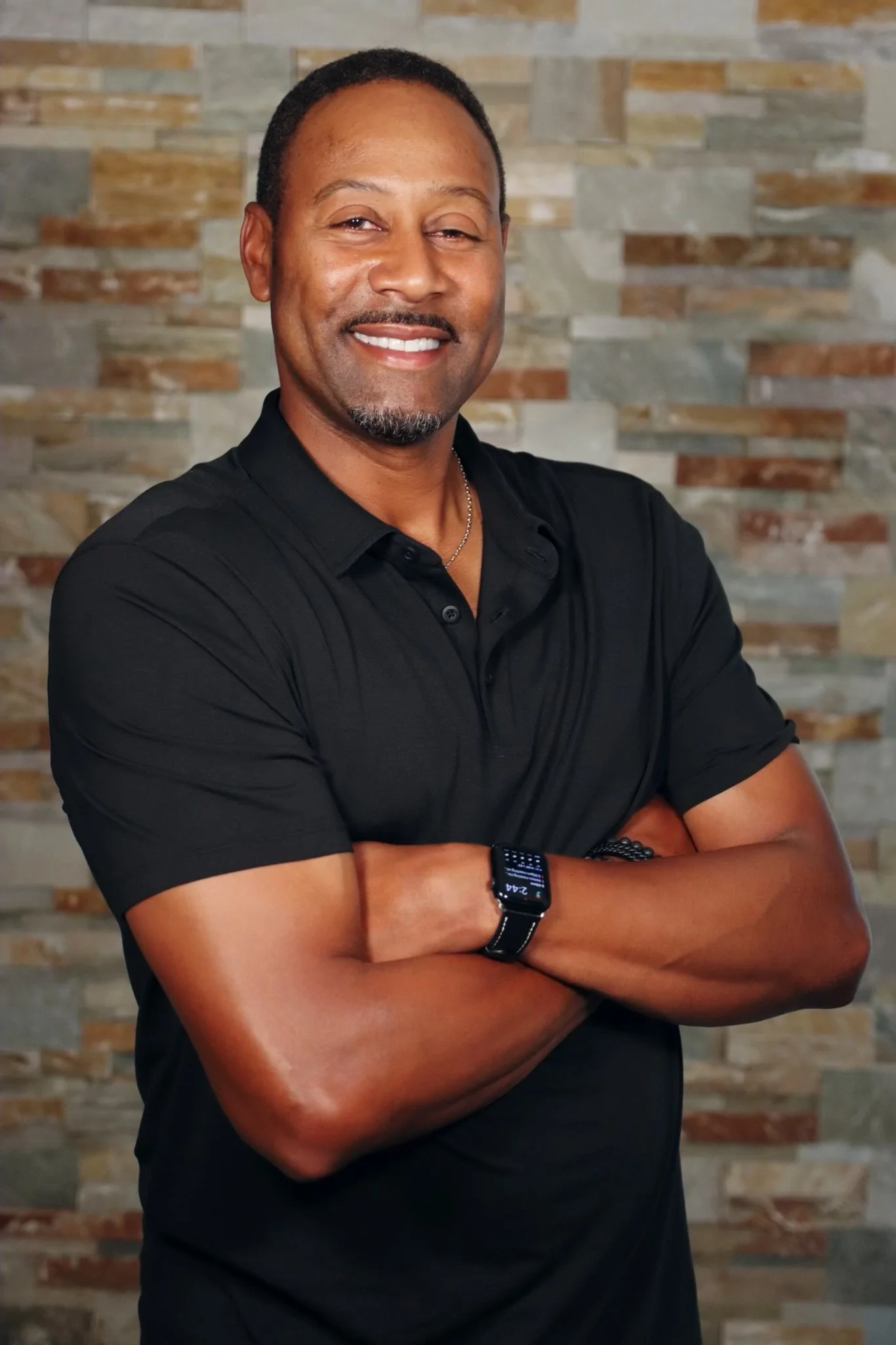 A man with short hair and a goatee smiling, crossing his arms, wearing a black polo shirt and a smartwatch, standing in front of a textured stone wall.