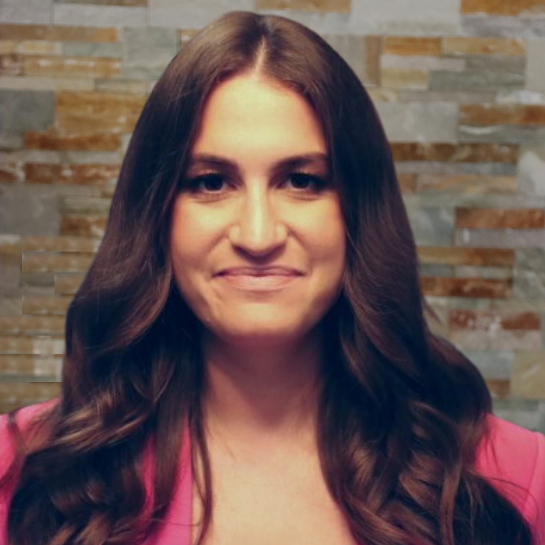 A woman with long, wavy brown hair smiling at the camera, wearing a pink top, standing in front of a stone wall background.