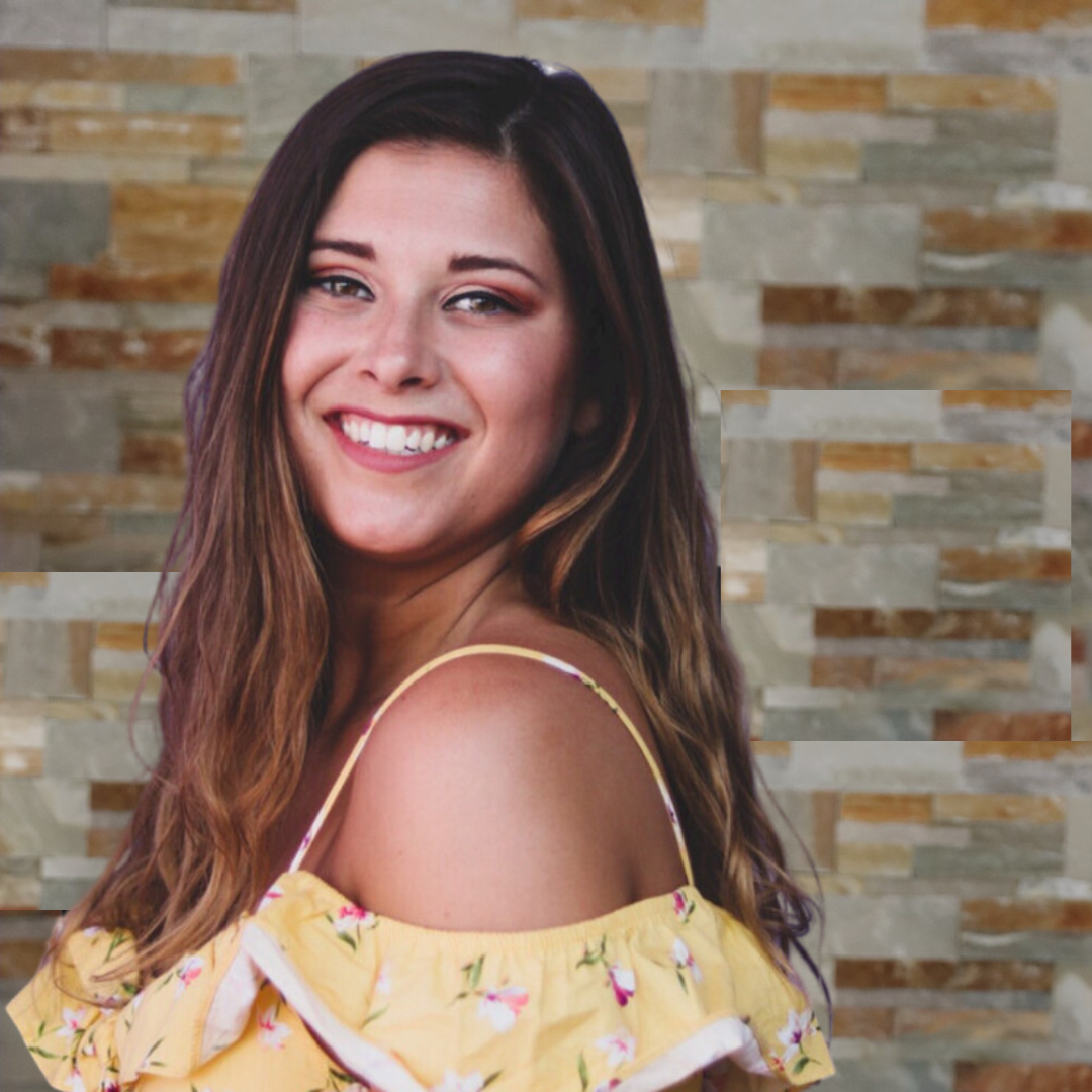 Smiling young woman with long brown hair, wearing a yellow floral off-shoulder top, standing in front of a stone wall background.