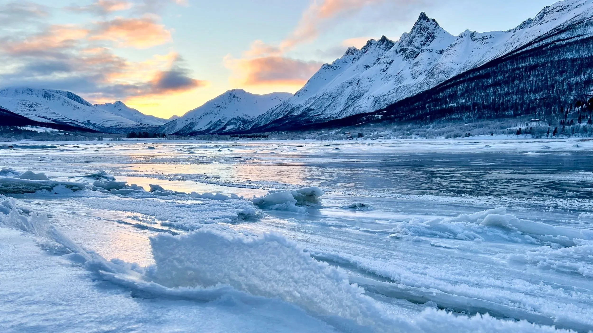 Snow-covered frozen lake surrounded by snow-capped mountains during sunset with colorful clouds in the sky.