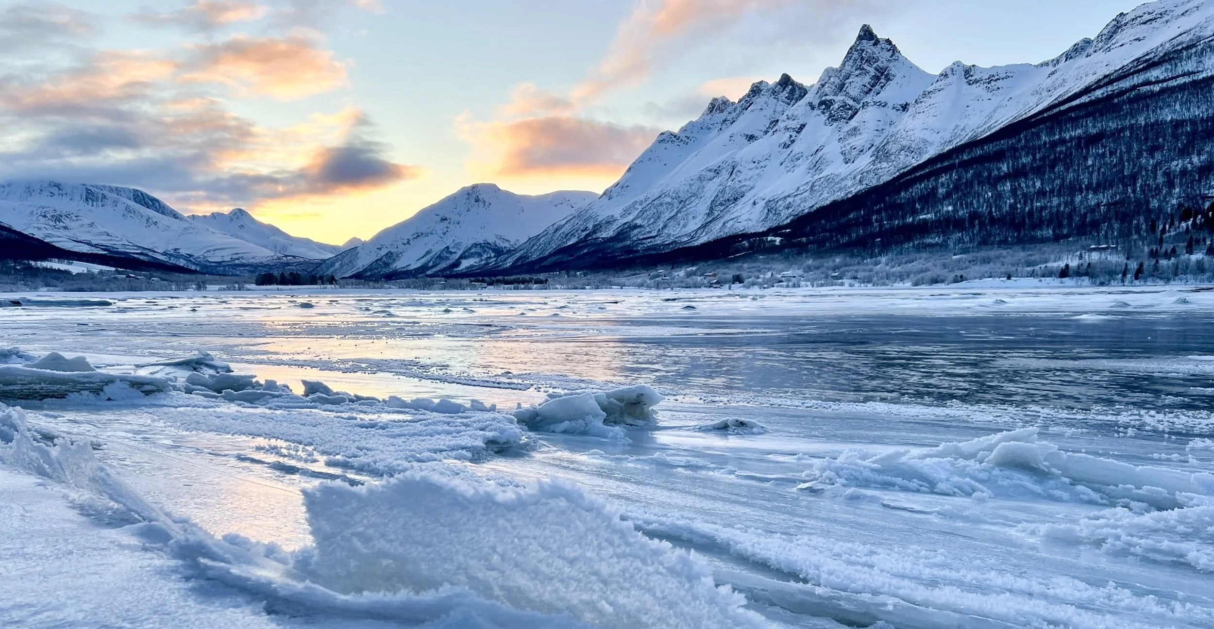 Snow-covered frozen lake with mountain range in the background during sunset, with clouds and a colorful sky.
