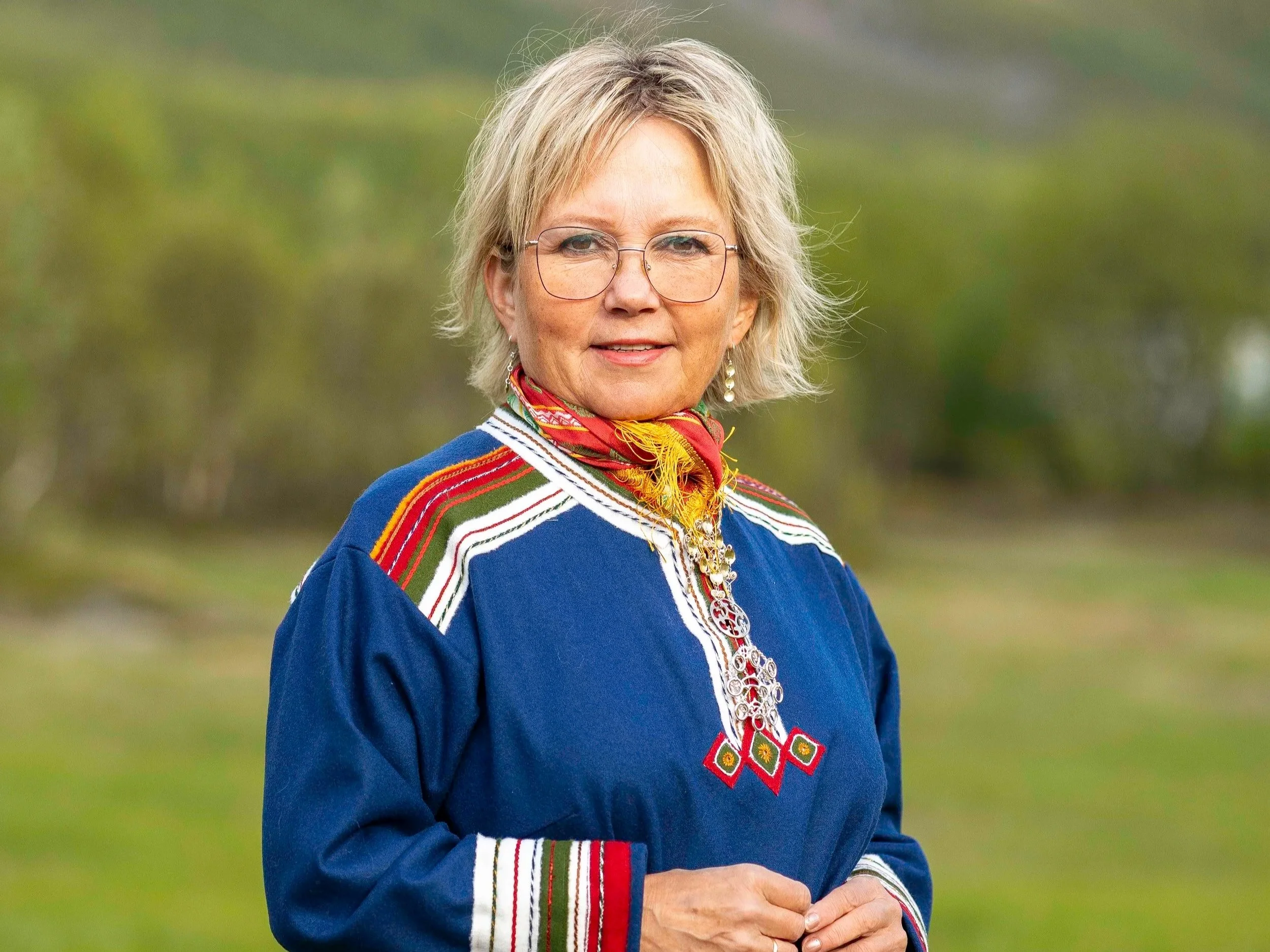 A woman in traditional Sami clothing standing on a rocky beach with water and mountains in the background.