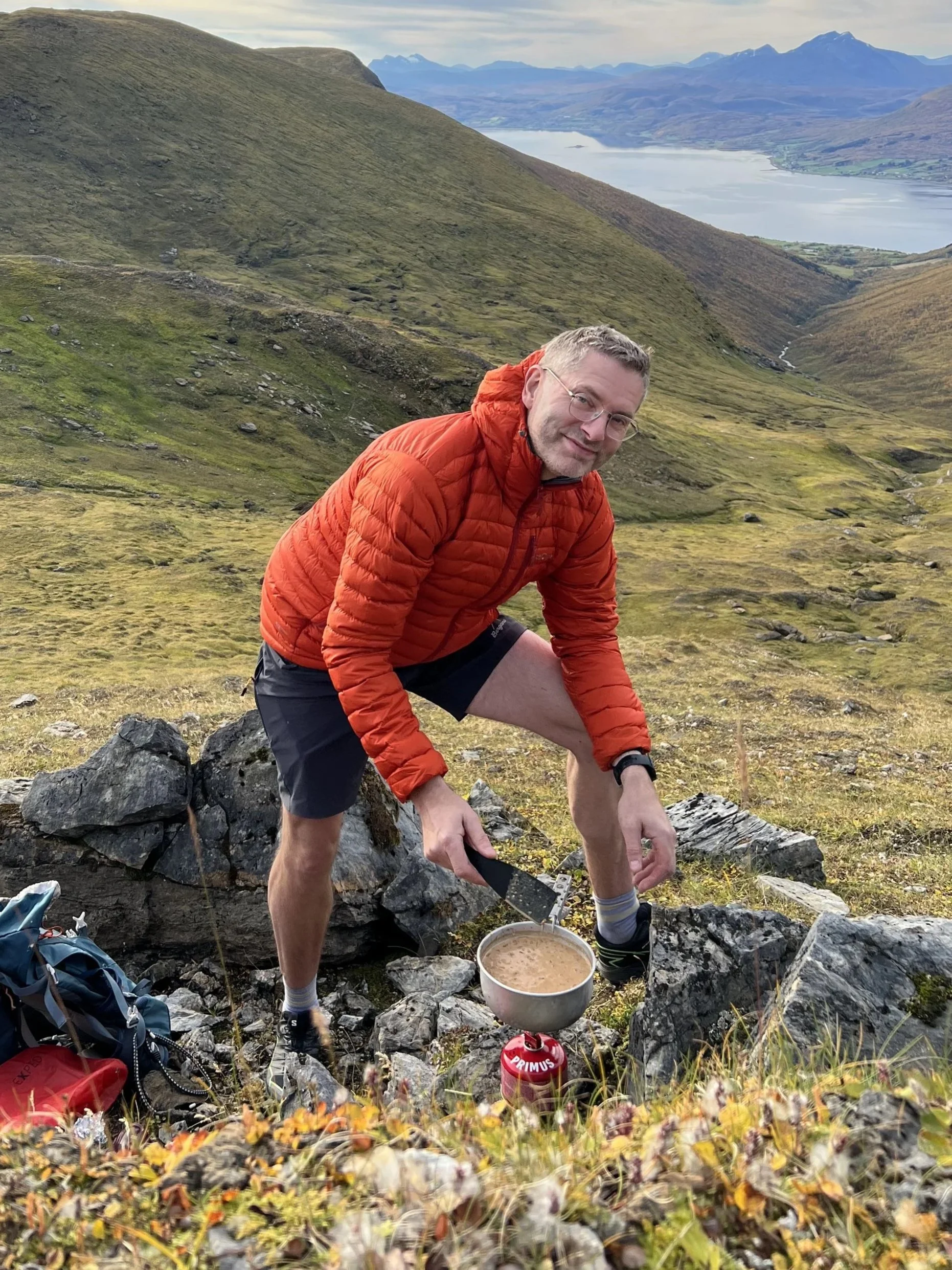 A man in outdoor clothing, including an orange jacket and shorts, crouches on rocky terrain in a mountainous landscape with green hills and a lake in the distance. He is preparing food using a light-colored pot over a camp stove, with backpack and gear nearby.