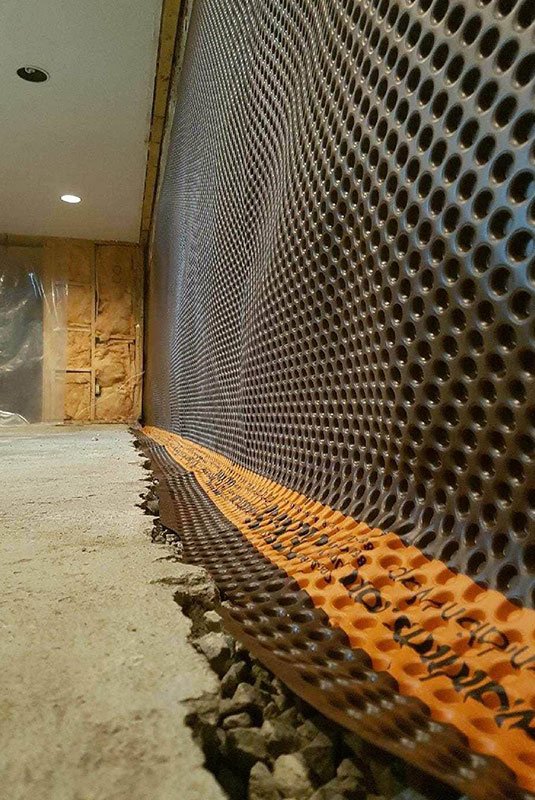 Construction site showing a perforated metal wall, orange safety tape, and unfinished flooring with scattered gravel and debris.