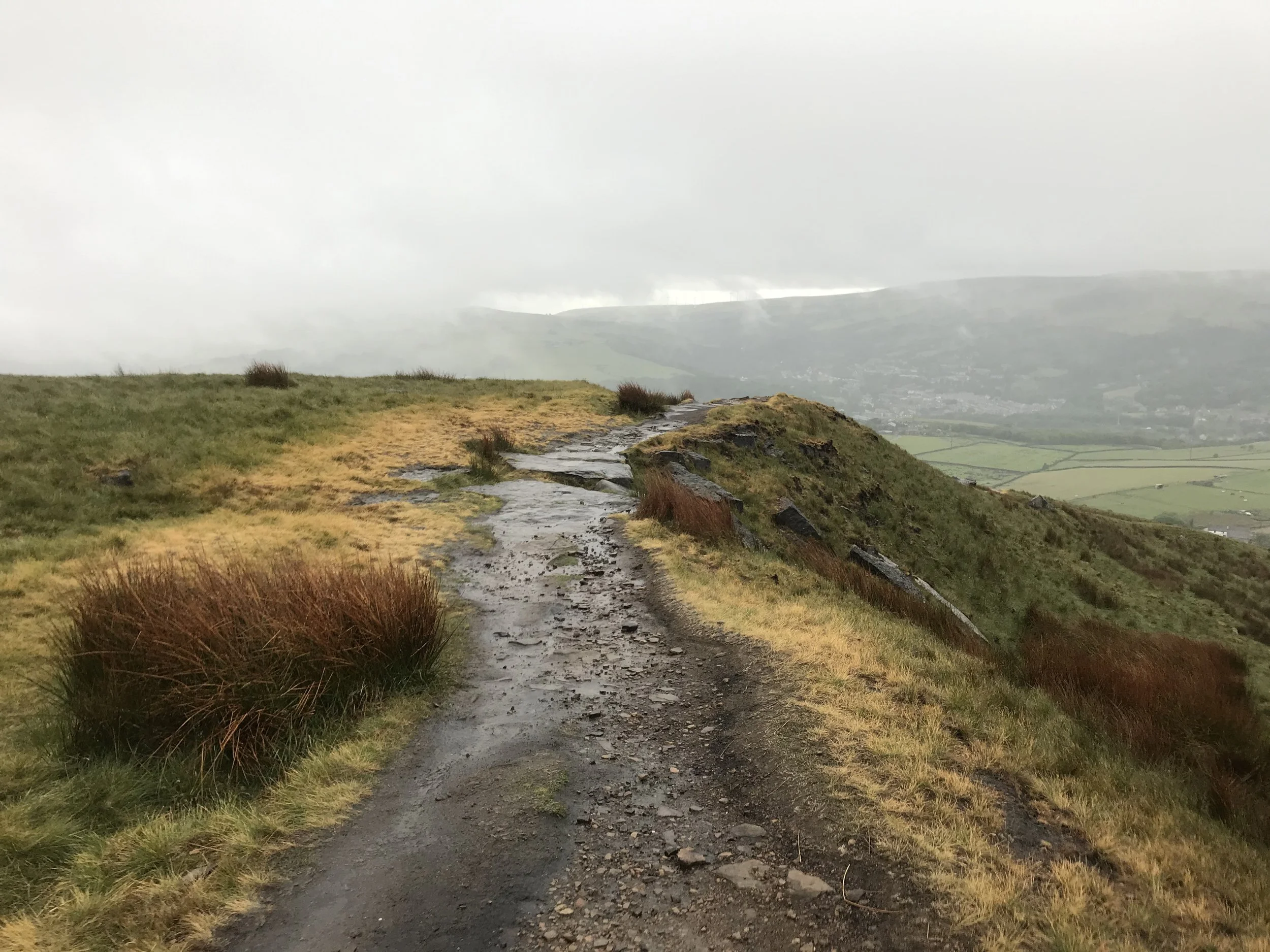 A muddy trail winding along a grassy hillside overlooking a distant valley with fields and scattered buildings. Overcast sky with low clouds.