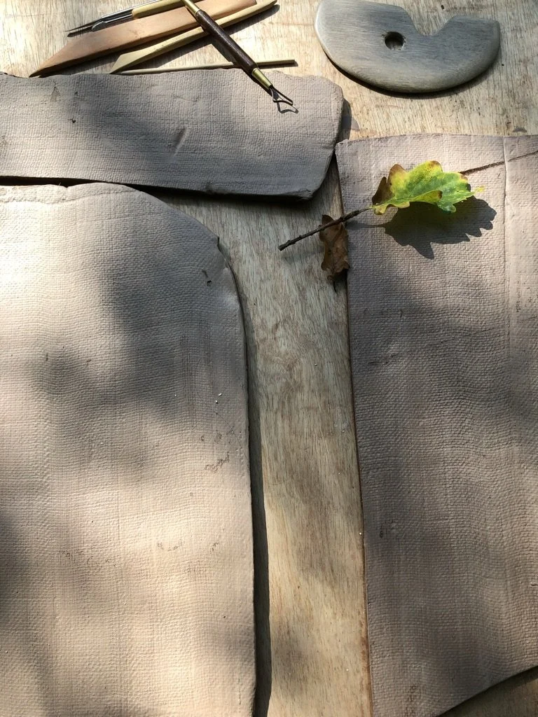 A crafting scene with clay pieces, sculpting tools, a clay modeling tool, and a small branch with leaves on a wooden surface.