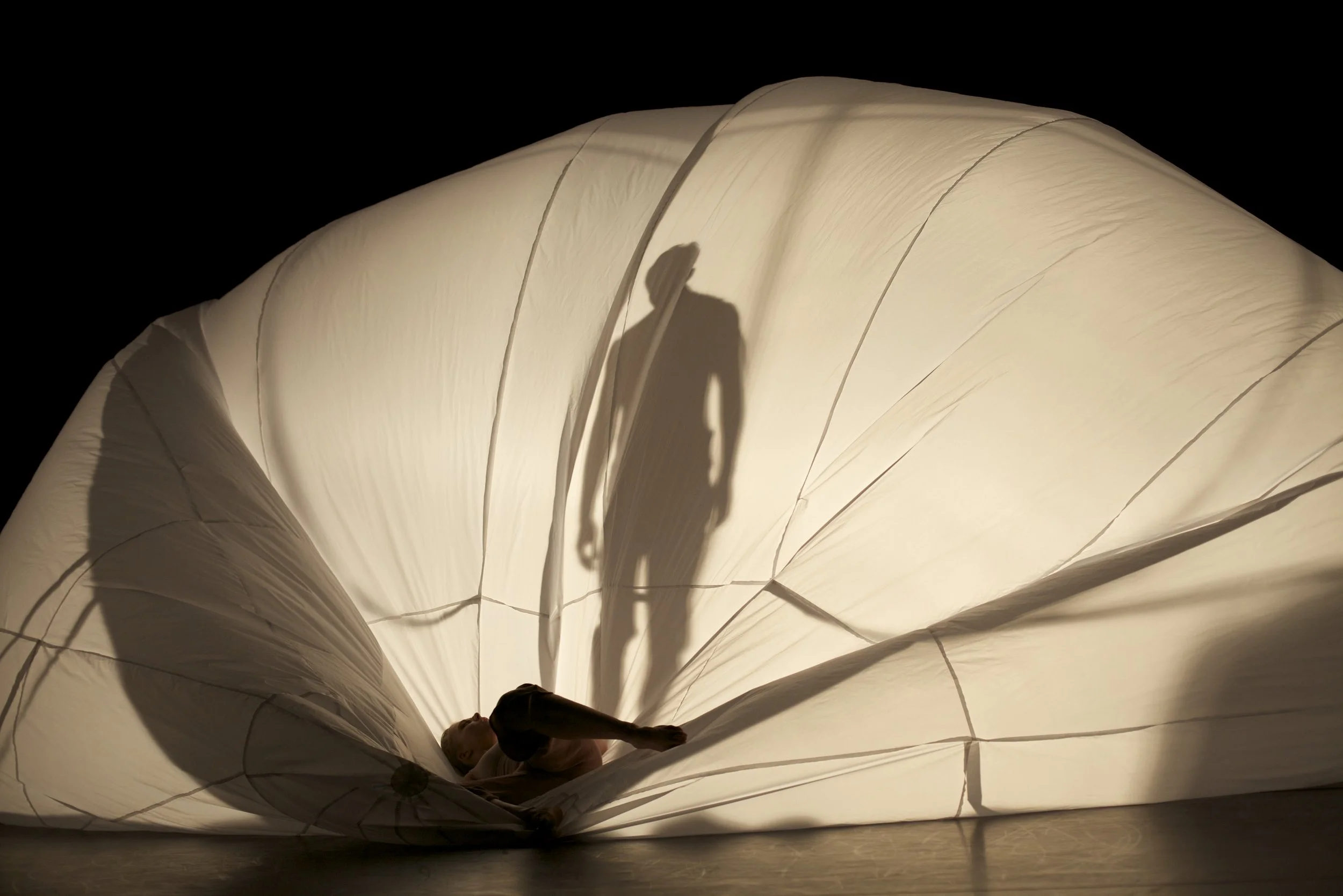 A performer lying on stage next to a large, illuminated fabric sculpture with a shadow of a standing person projected onto it.