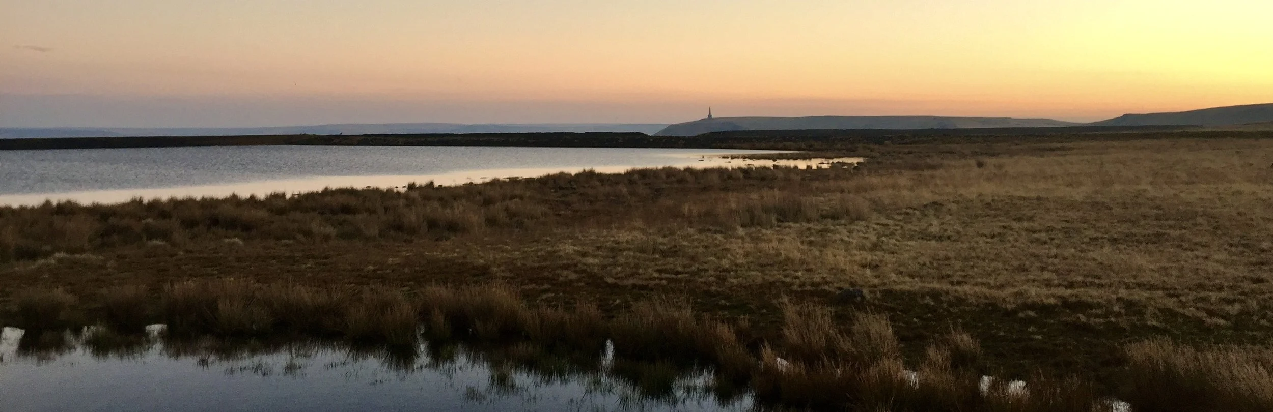 A tranquil landscape at sunset showing a body of water, grassy and marshy land, and distant hills with a monument or tower on top.