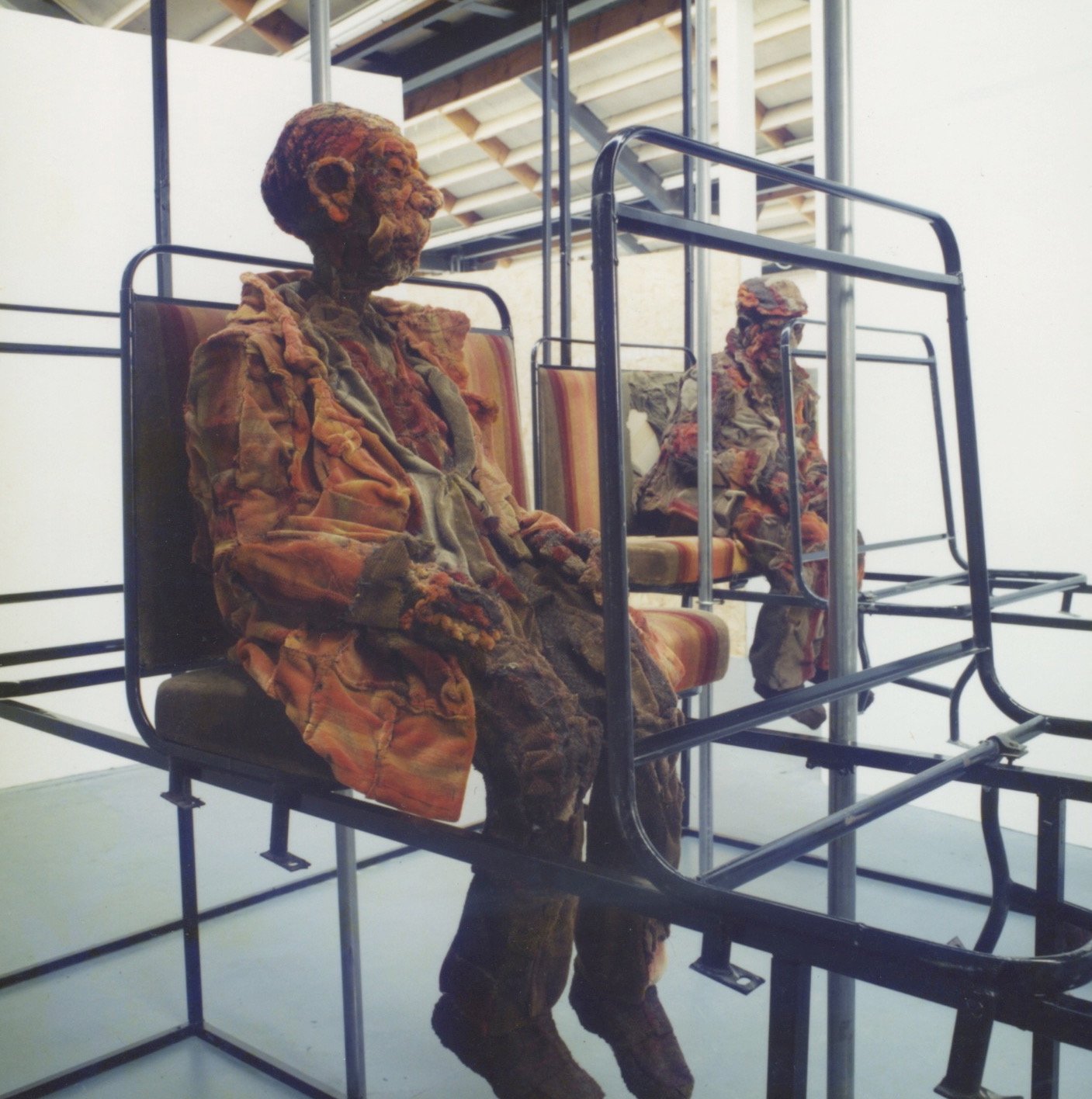 Two mannequins dressed in patchwork clothing, seated on metal frames with striped cushions in an art installation or museum setting.