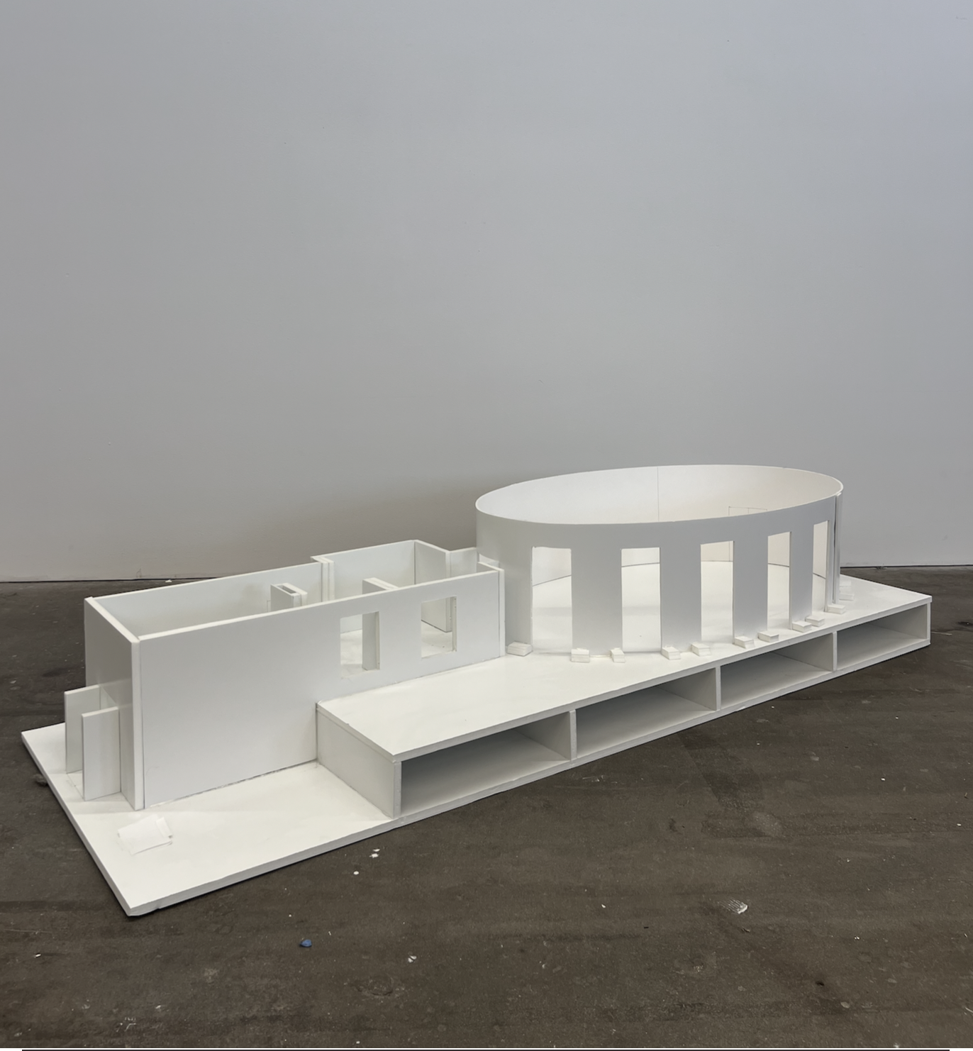 White architectural scale model of a building with curved and rectangular sections displayed on a dark floor against a white wall.