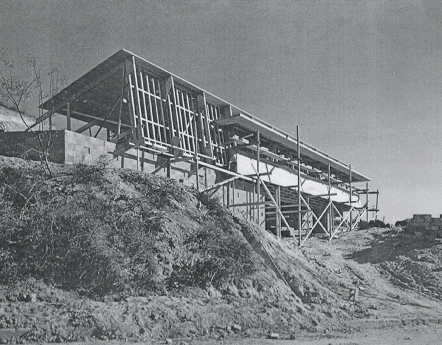 Black and white photo of a house under construction on a hillside, with wooden framing and scaffolding.