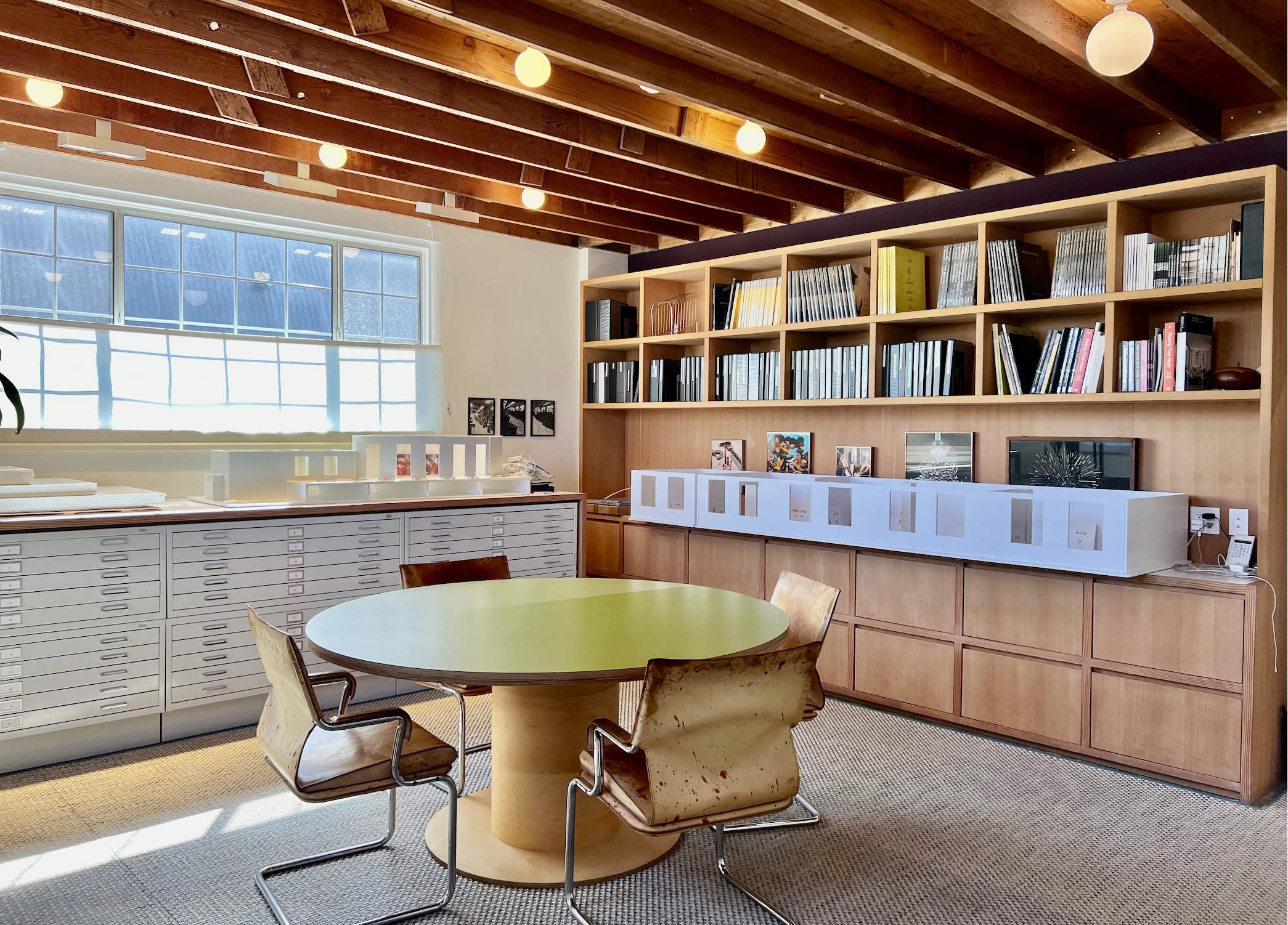 Office meeting room with round wooden table, beige chairs, built-in wooden bookshelves filled with books and framed photos, large window with sunlight, wooden ceiling beams, and a row of white display cases on the right side.