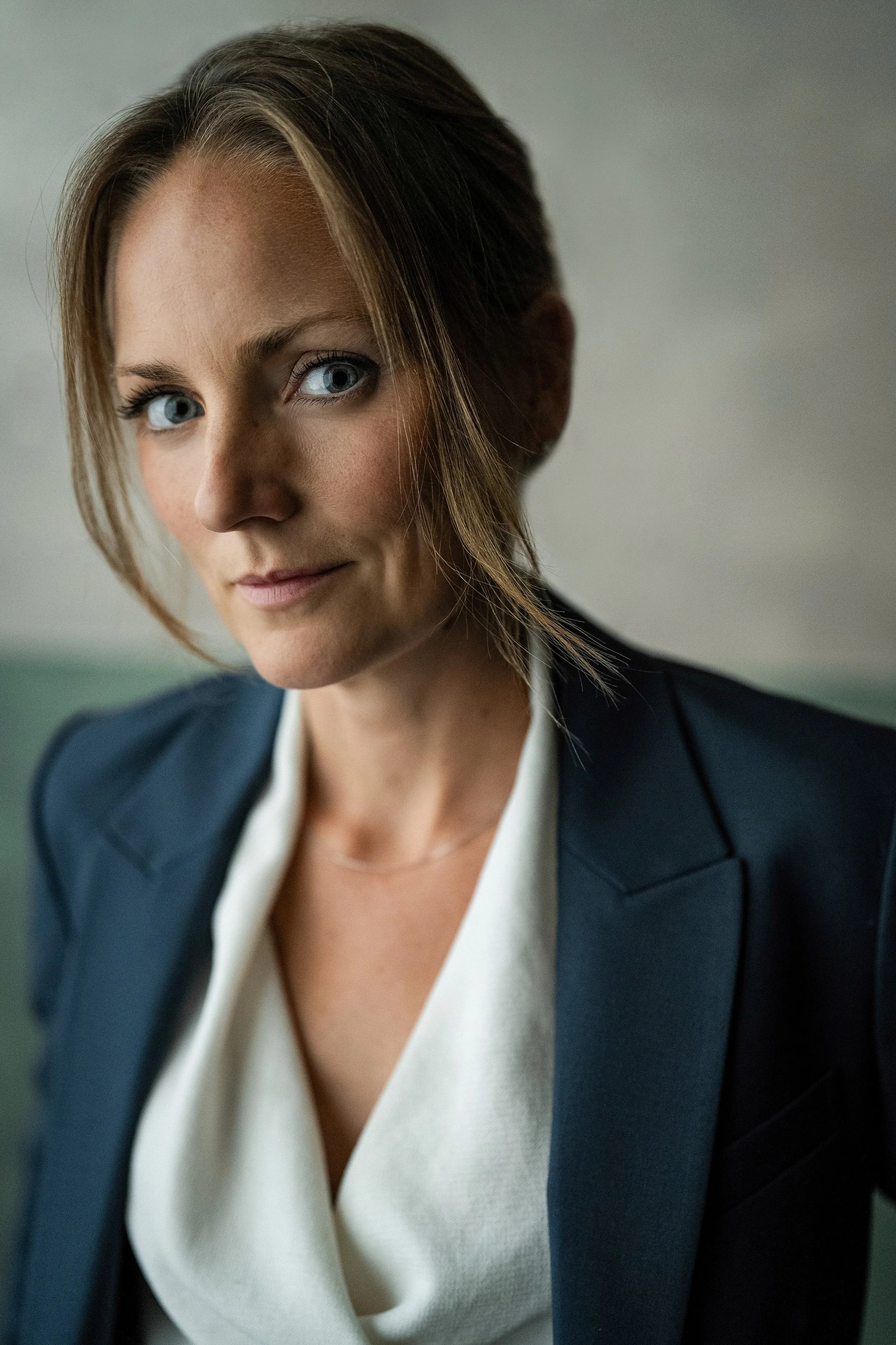 Caucassion woman with blonde hair and blue eyes and a petrol suit with a white blouse.