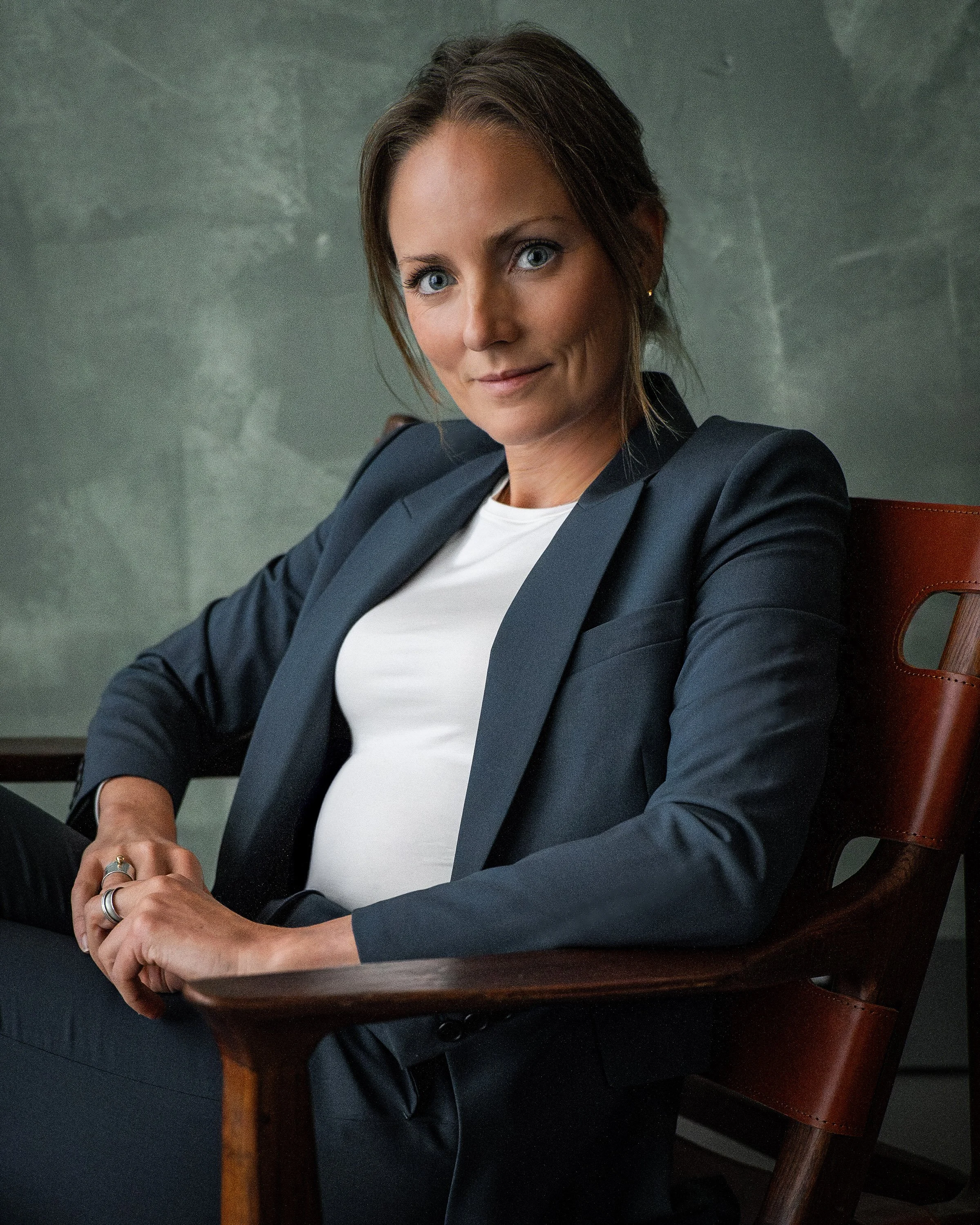 Businesswoman with brown hair and blue eyes sitting on a wooden chair, wearing a black blazer and white shirt, with a confident expression.