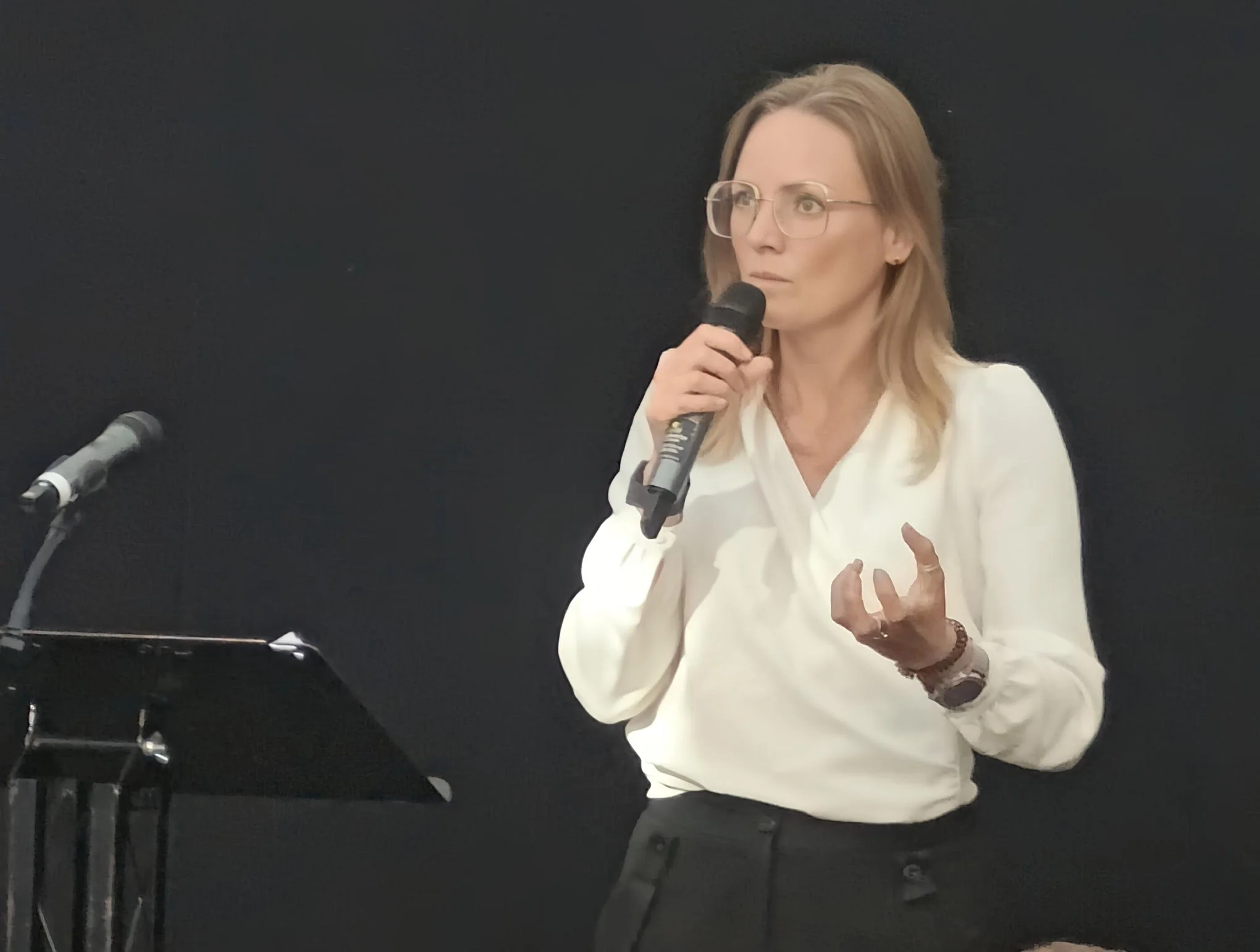 A woman with blonde hair, glasses, and wearing a white blouse, standing against a black background, holding a microphone and gesturing with her other hand while speaking.
