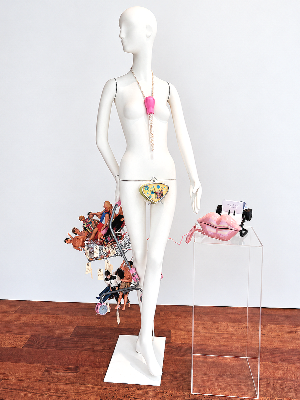 "HelloHoney" September 2026. Seeking male DNA for a pregnancy takes many possible forms. This mannequin holds a toy shopping cart full of Ken dolls, each tagged with pertinent information. Some are anonymous sperm donors from a bank, some are acquain