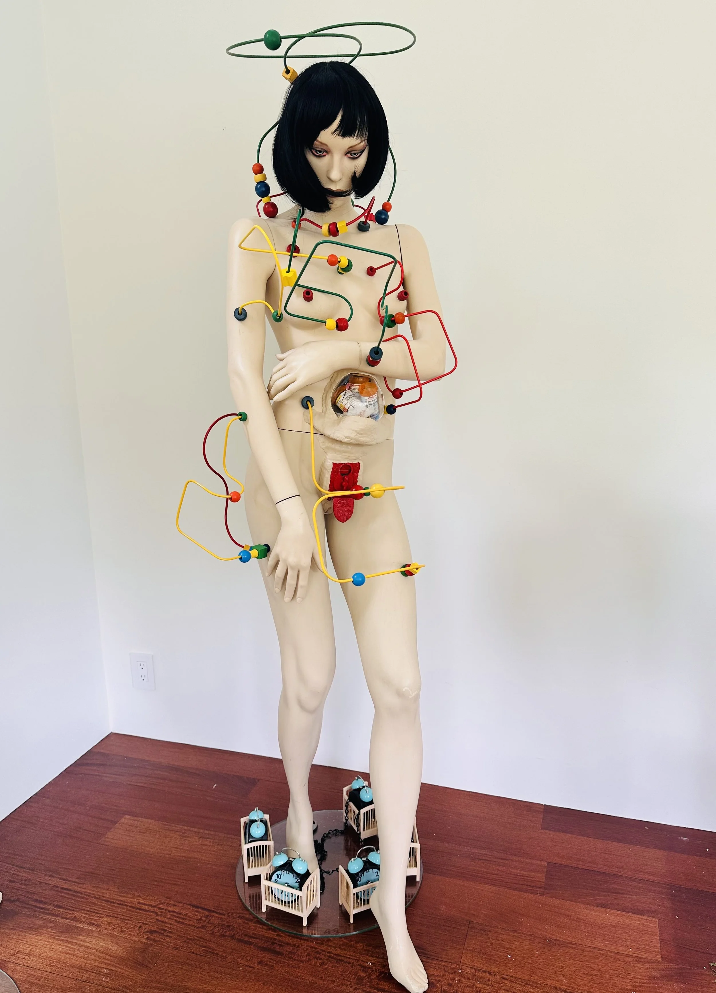 PostpartumAnxiety, 2026. This vintage mannequin is caged in by the heavy wires of vintage bead mazes. They pierce her arms, legs, and torso. Her gum ball machine contains the artist's empty prescription bottles. Her feet are chained to a series of cr