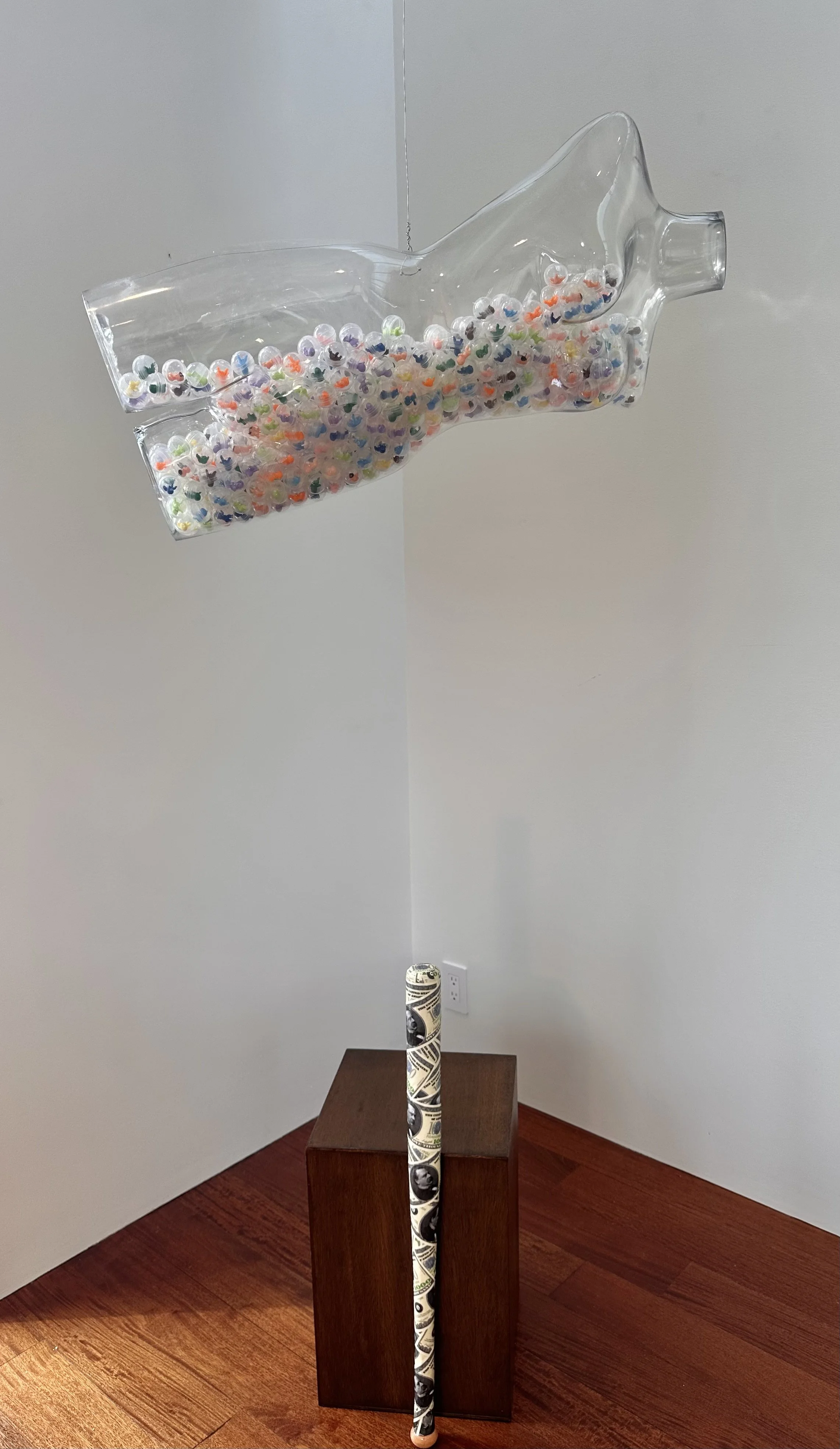 At Any Cost 2026. Piñata and bat. A transparent mannequin contains used toy vending capsules holding tiny colorful babies. The bat is découpaged with (faux) $1000 bills. The piñata game represents anticipation and hope. This piece points to the dream