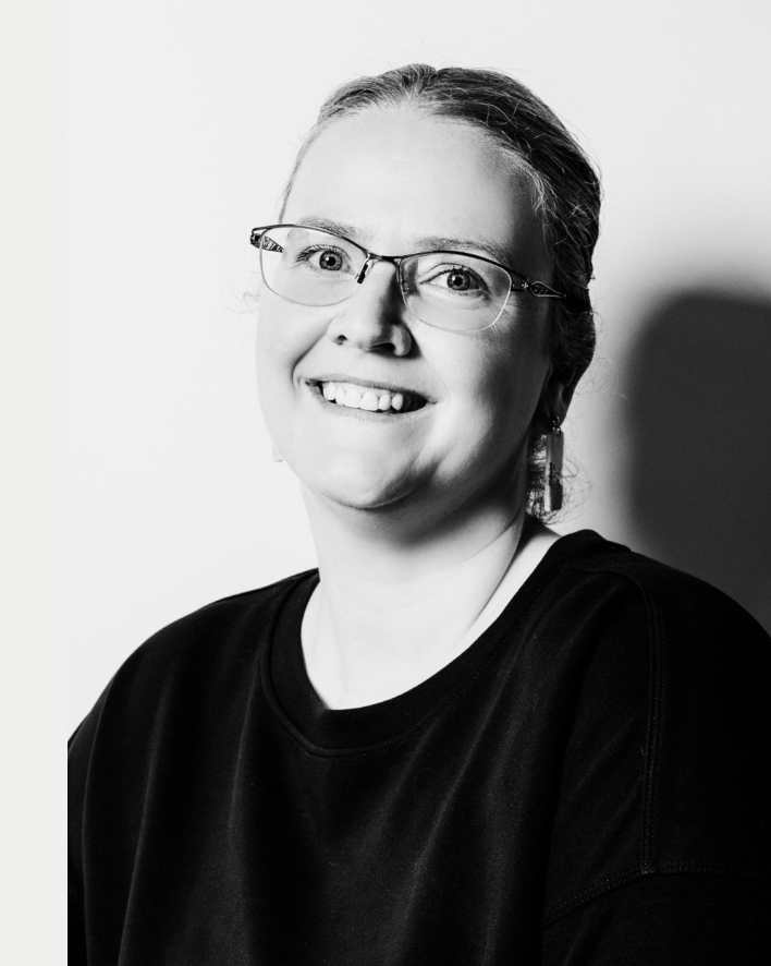 Black and white portrait of a woman smiling, wearing glasses and earrings, with her hair pulled back, in front of a plain background.
