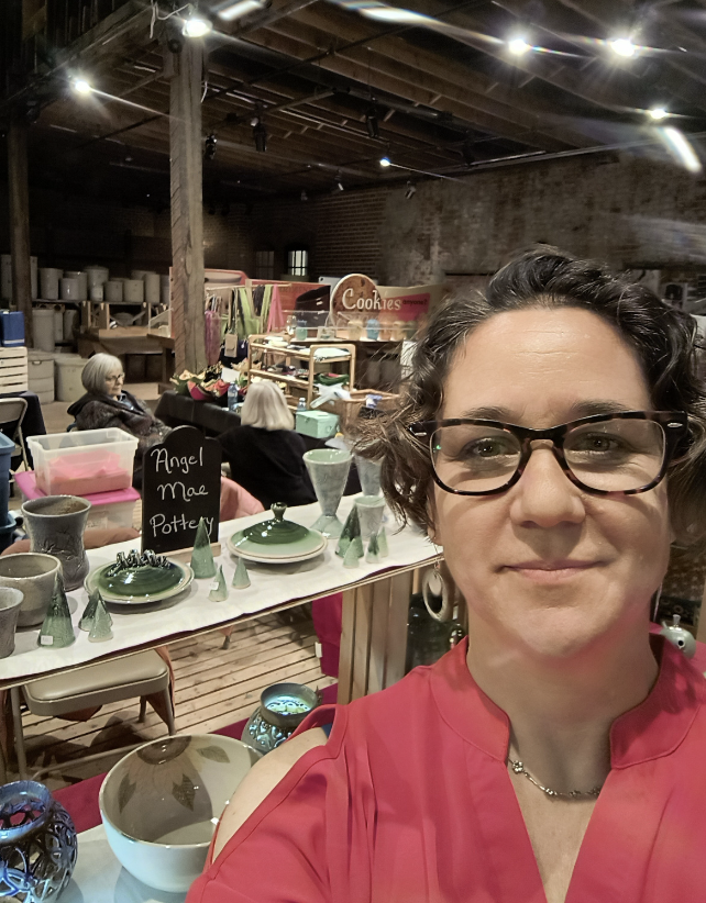 Selfie of a woman with short curly hair and glasses in a red top, indoor craft fair with pottery and home decor items, some people browsing in the background.